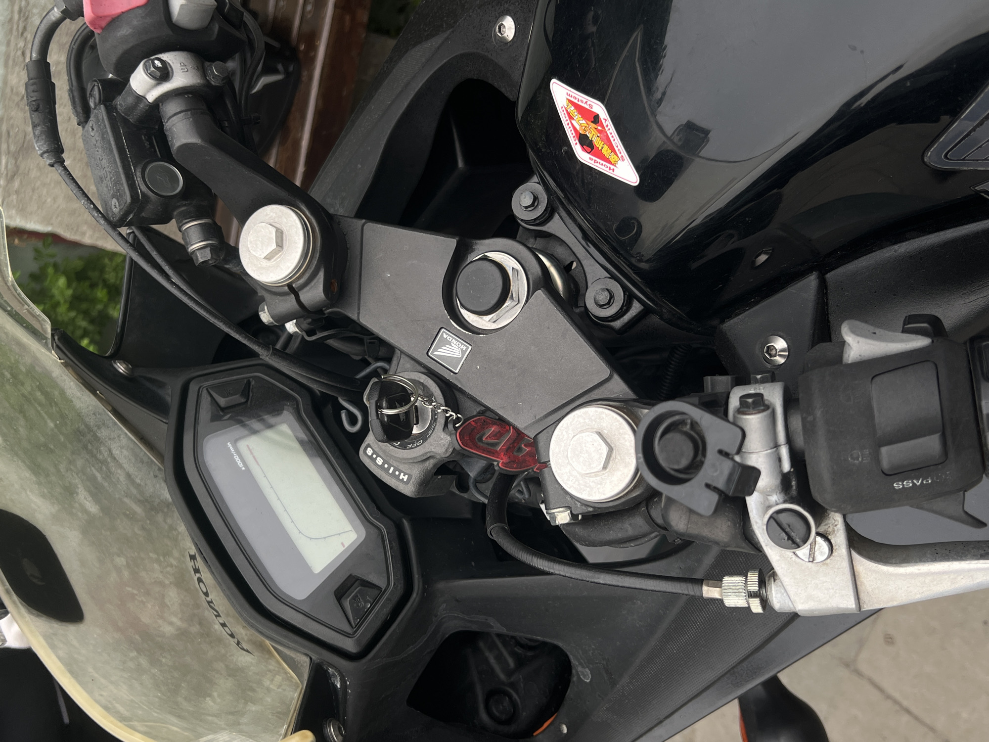 The image shows the front portion of a black Honda motorcycle, likely a sportbike model. The viewpoint is from above the handlebars, looking down. Key elements include the motorcycle's ignition switch with a key inserted, labeled "H・I・S・S" (Honda Ignition Security System), the dashboard instrument panel displaying the tachometer with the Honda logo, and the steering assembly with visible bolt heads. A sticker, presumably for the Honda Ignition Security System, is affixed to the fuel tank. The brake lever and associated controls are also visible. The image does not contain any indications of a location in the city of Horodok, Ukraine.