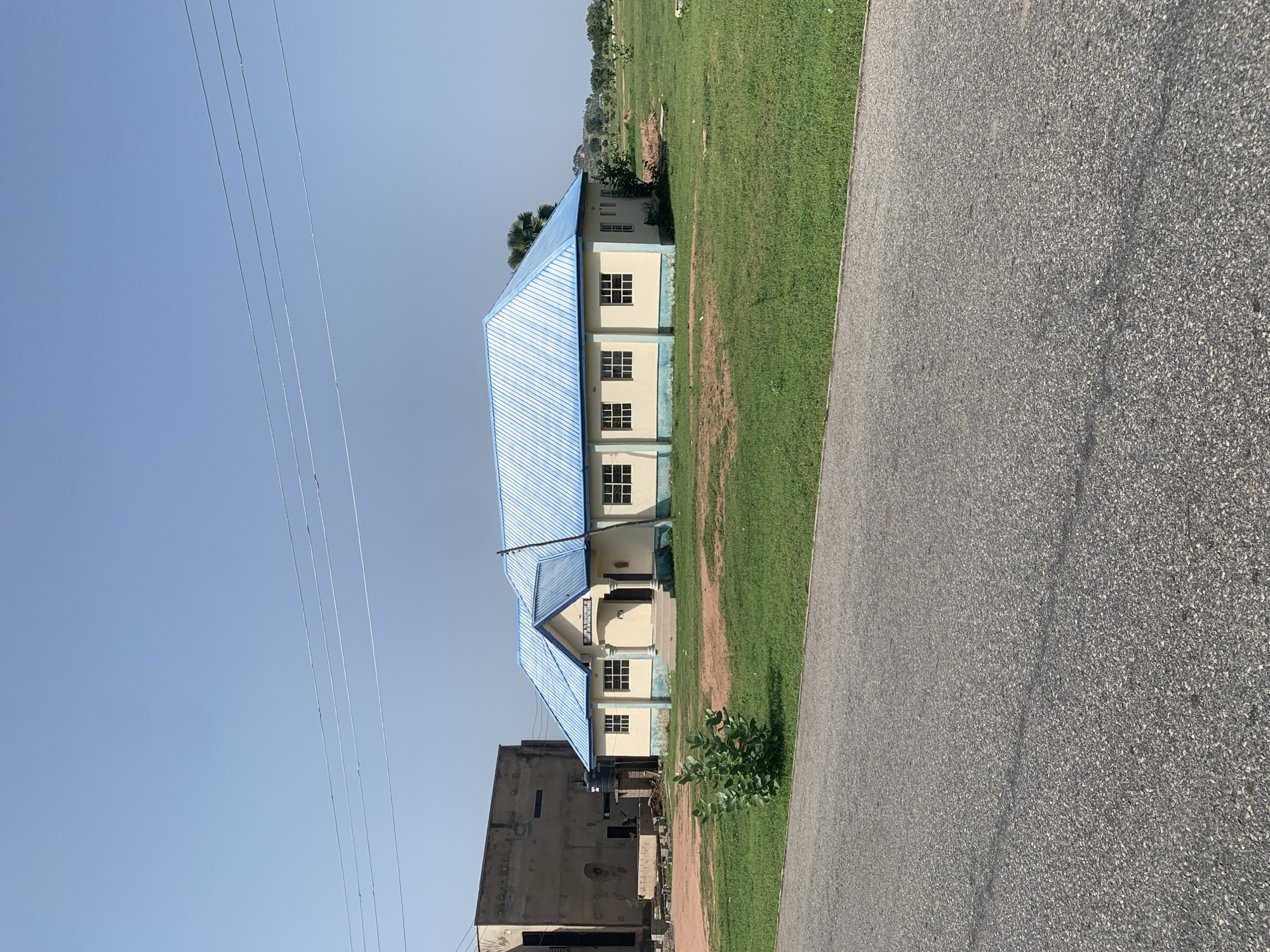 This is a full, eye-level, exterior shot of a single-story building with a light blue metal roof. The building is painted a pale yellow with a light blue trim. It has a symmetrical facade with a central entrance and several evenly spaced windows on each side. The windows appear to be multi-paned with dark frames. A small green bush is in the foreground to the left of the building. To the left of the building is a taller, unfinished concrete structure. The scene is set in a grassy, open field under a clear blue sky. Power lines are visible in the upper portion of the image. The building appears to be the "Jalingo Local Education Authority Primary School".