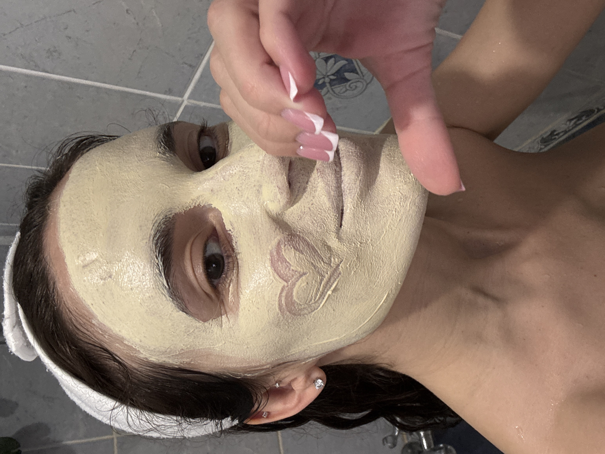 A woman lies supine in a bathroom. Her face is covered with a light yellow facial mask, with a heart drawn on her left cheek. A woman's hand with pink and white manicured nails is positioned above the woman's face. The woman is wearing a white headband, and two earrings are visible in her left ear. The bathroom has grey tiled walls and blue accent tiling. The location cannot be determined from the image.