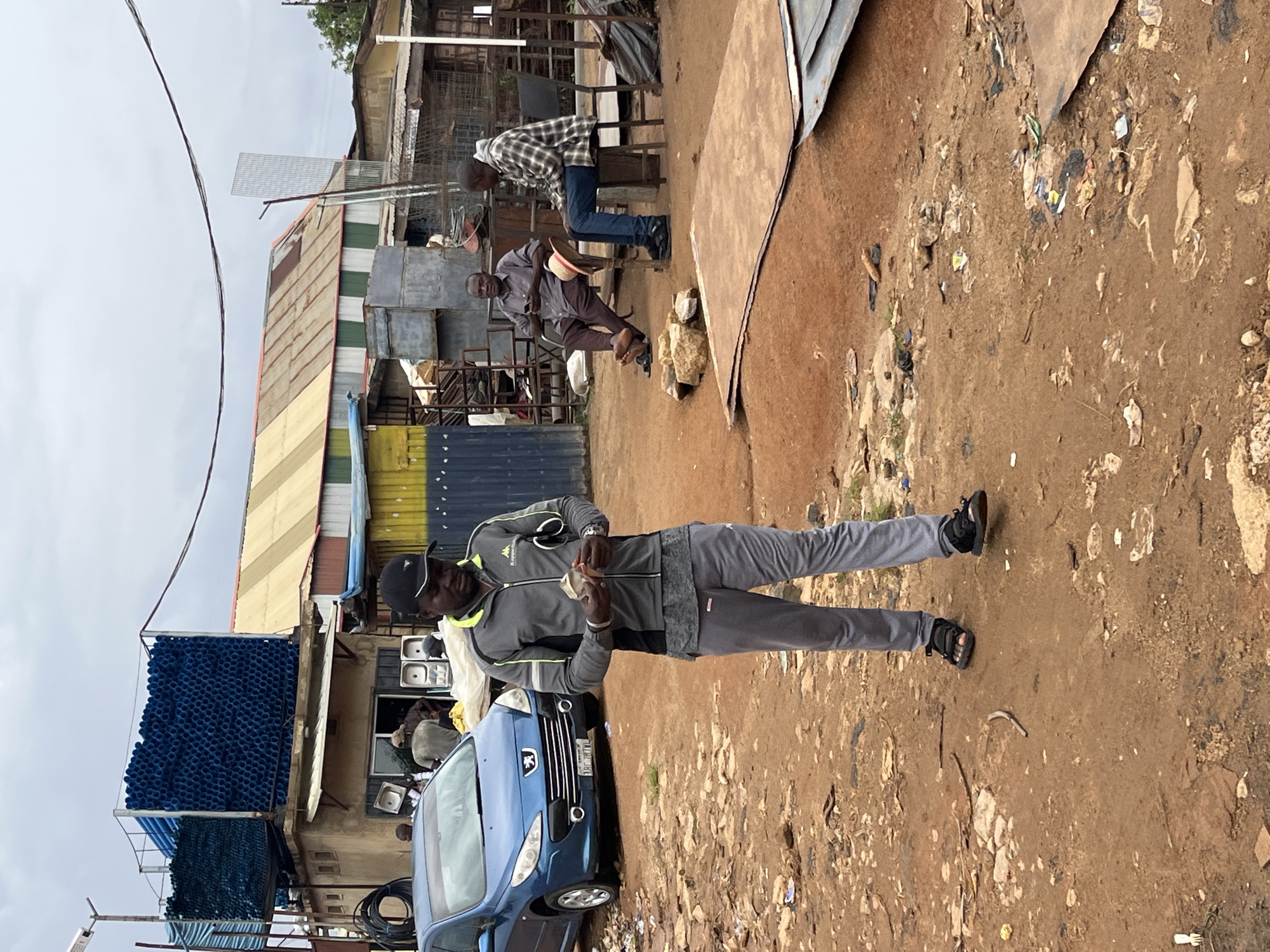 The image depicts a scene in a bustling, outdoor marketplace in Gura Top, Nigeria. In the foreground, a man wearing a grey athletic jacket and grey sweatpants is holding some money in his hands. Behind him, a blue hatchback car is parked. Further back, two other men are seated; one in a light brown shirt and trousers, and the other in a plaid shirt and blue jeans. The environment is a dusty, unpaved area with various structures, including a building with a corrugated metal roof, stacks of blue pipes, and other stored materials. The overall atmosphere suggests a busy commercial hub.