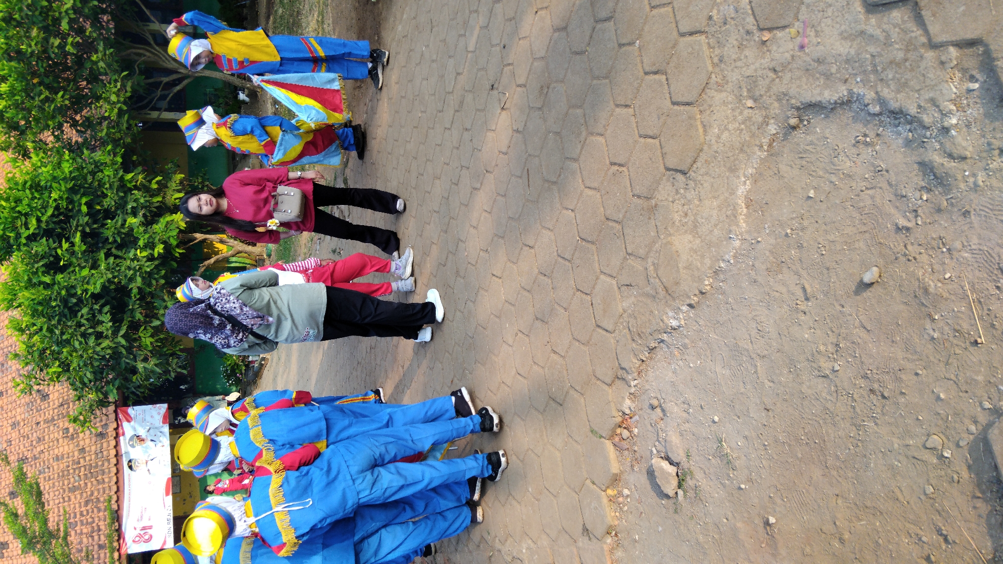The image captures a group of children in marching band uniforms, a woman, and another individual standing on a paved area with hexagonal tiles. The children, dressed in bright blue uniforms with yellow accents and hats, appear to be either preparing for or have just finished a performance. One child is holding a colorful flag. The woman in the foreground is wearing a dark pink shirt and black pants, with a beige purse slung across her shoulder. She is looking towards the children. The environment suggests an outdoor setting, possibly a schoolyard or a public space in the city of Sembojo, Indonesia, with trees and a building with a tiled roof visible in the background. The ground is a mix of paved tiles and dirt. The lighting indicates it is daytime, likely late afternoon due to the warm tones.