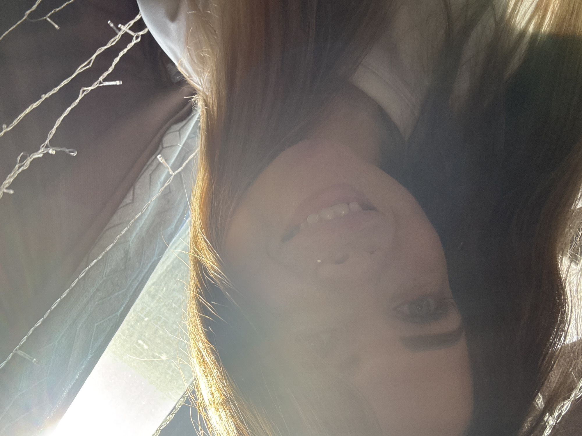 A bright, upside-down portrait captures a smiling person, likely a young woman, with long, light brown hair. The person's face is positioned towards the bottom of the frame, tilted, and they are looking towards the viewer with a wide smile that reveals their teeth. A significant lens flare and areas of overexposure dominate the left side of the image, originating from a strong light source, likely sunlight, which brightly illuminates the scene and highlights strands of hair.
In the background, decorative string lights with small bulbs are visible in the upper left quadrant. Partially obscured by the bright light and the person's hair, there is also some dark-colored fabric, possibly a curtain or upholstery. A glimpse of a white garment, perhaps a collar, can be seen near the person's shoulder. The overall impression is one of a playful or cheerful moment, captured indoors during daytime. The unusual upside-down orientation contributes to an informal and candid aesthetic. No text is visible in the image.