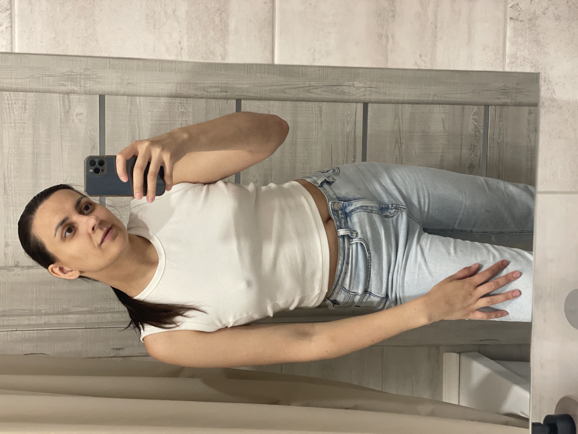 A woman is taking a selfie in a bathroom. She is wearing a white t-shirt and light blue jeans. She has dark hair pulled back in a ponytail. The background consists of gray wood-paneled walls and a white tiled wall. The location is an indoor bathroom setting. The city is Unknown, Unknown.