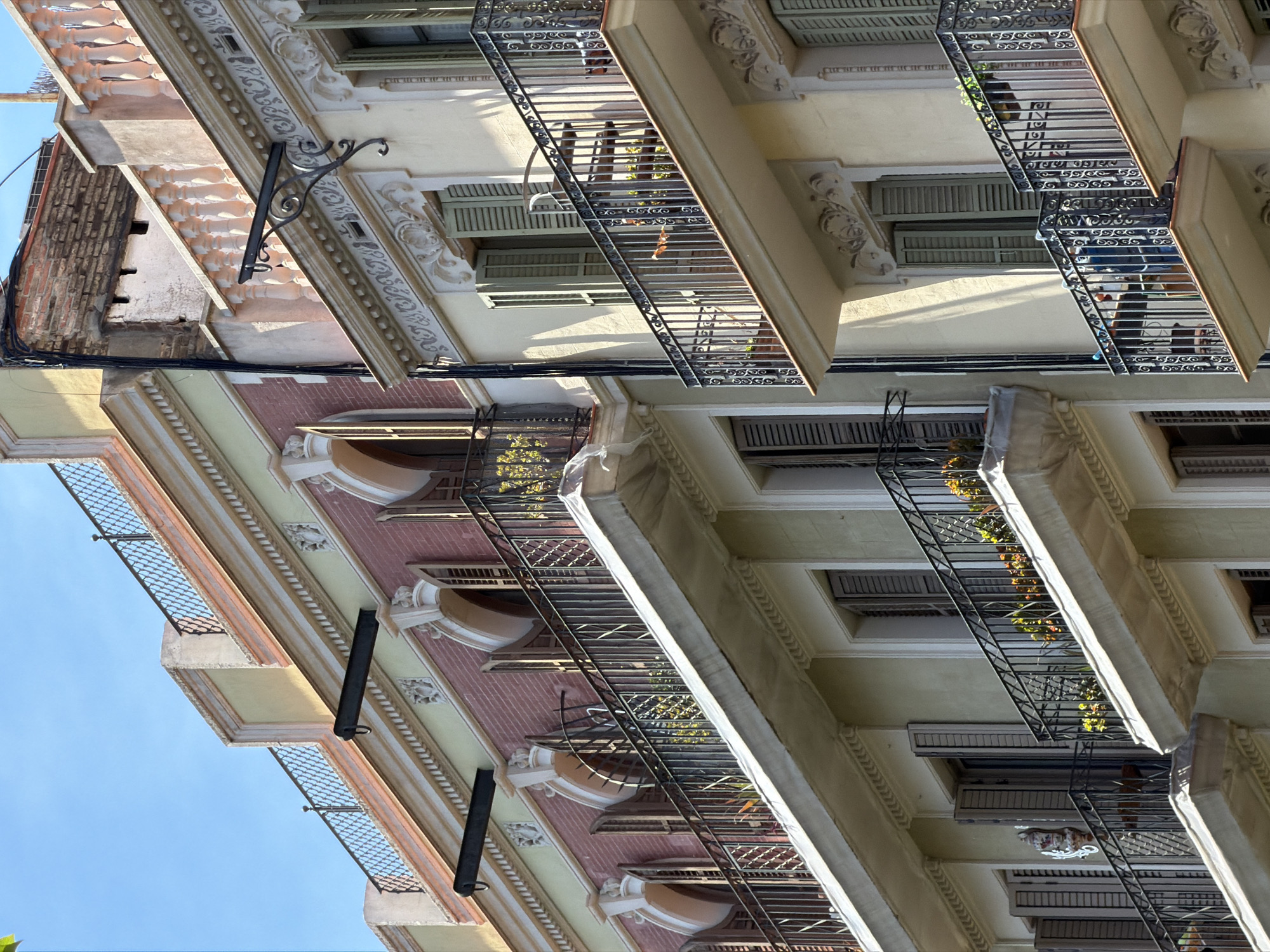 An upward angle shot shows the facades of two connected buildings in Barcelona, Spain, under a clear blue sky. The buildings feature ornate architectural elements, including arched windows, decorative cornices, and wrought-iron balconies filled with plants. The building on the left has a pink brick lower section and a yellow upper section, while the building on the right is predominantly cream-colored. Balconies have metal railings with intricate scrollwork and are furnished with potted plants. Some windows have closed wooden shutters, while others are open, revealing glimpses of the interiors or the outdoor space. The scene captures the architectural style of Barcelona, characterized by its blend of color, detail, and residential design.