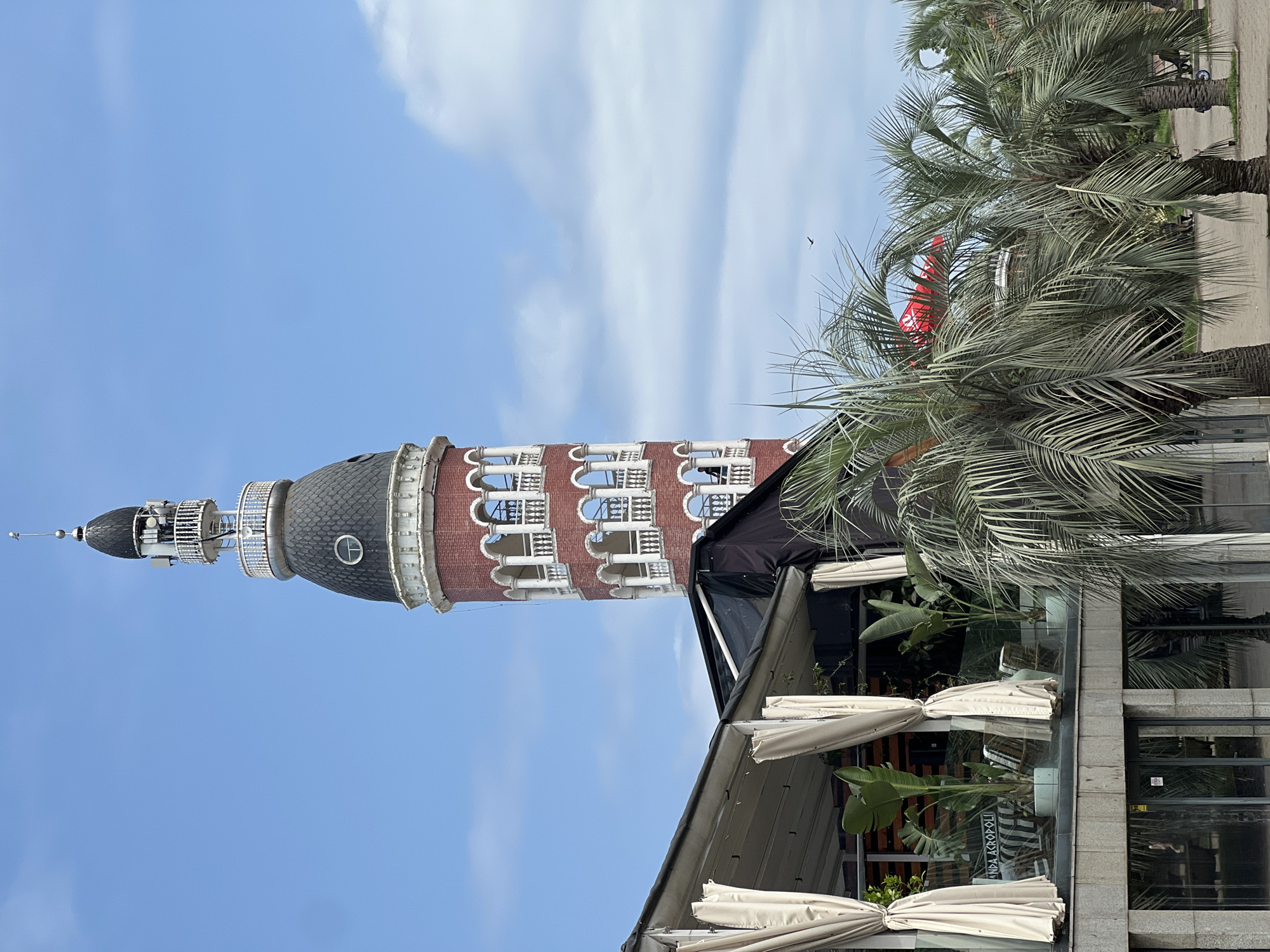 The image captures the Alphabet Tower in Batumi, Georgia, a landmark located along the Batumi Boulevard. In the foreground, a modern building with white curtains and lush green plants is visible. Beyond this, the distinctive red brick Alphabet Tower rises against a clear blue sky with scattered clouds. The tower features multiple levels with arched openings and white balustrades. Palm trees frame the scene, suggesting a coastal or tropical environment. A red banner or flag is partially visible behind the palm trees in the mid-ground. The overall impression is of a bright, sunny day in a tourist-oriented area.
