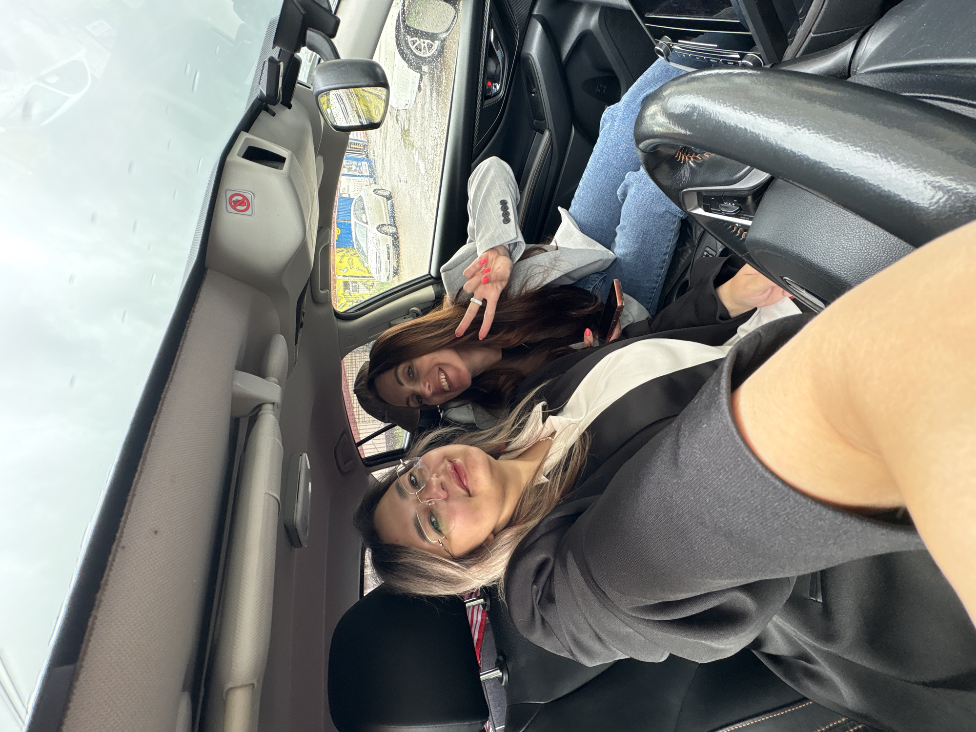 The image is a vertical selfie taken inside a car, featuring two women in Birobidzhan, Russia. The woman in the foreground, likely holding the camera, has blonde-streaked hair, wears glasses, a black blazer over a white top, and is smiling directly at the viewer. Her arm is visible on the right.
Seated behind her, the second woman with long, dark brown hair is also smiling and making a peace sign with her right hand. She is dressed in a light grey blazer and blue jeans, with a mobile phone in her left hand. The steering wheel is partially visible on the right side of the frame.
The setting is a car interior, with parts of the dashboard and rearview mirror visible. Through the windshield and side window, it appears to be a rainy or overcast day, with raindrops visible on the glass and blurred reflections of outdoor elements such as buildings and what appear to be other vehicles.
A distinct "no smoking" symbol—a red circle with a crossed-out cigarette icon—is visible on the car's interior panel near the windshield. Both women exhibit pleasant and happy expressions.