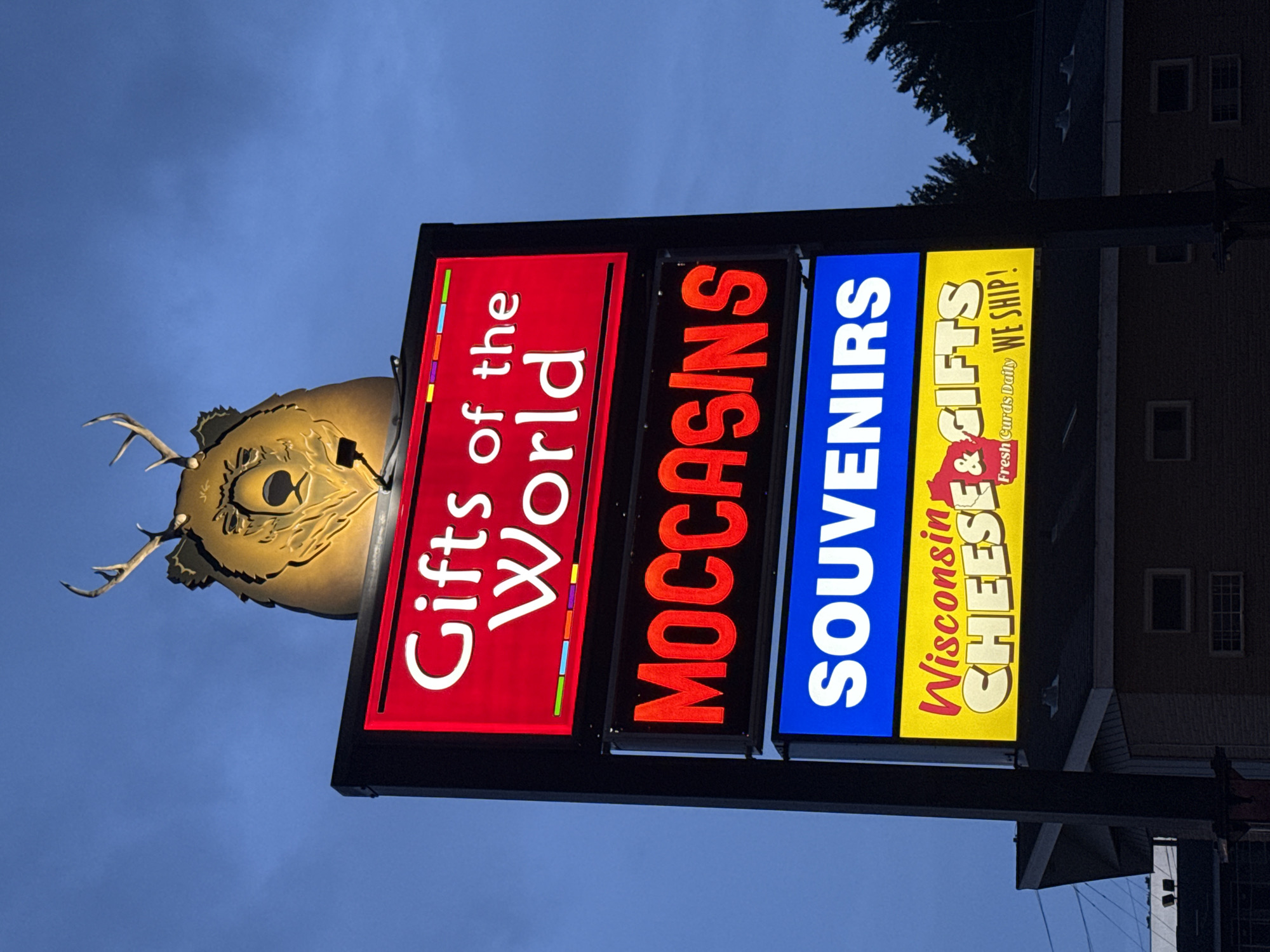 The image shows a lit-up sign at night in Lake Delton, Wisconsin, USA. The sign is mounted on a dark pole and has four sections. The top section features a carved wooden head of a bear with antlers, illuminated from within. Below the bear head, the sign has four illuminated panels. The top panel is red with white text that reads "Gifts of the World". The second panel is black with red text that reads "MOCCASINS". The third panel is blue with white text that reads "SOUVENIRS". The bottom panel is yellow with red and white text that reads "Wisconsin CHEESE & GIFTS Fresh Curds Daily WE SHIP!". The background is a dark blue sky with some tree branches visible on the right side. The sign is positioned in front of a building.