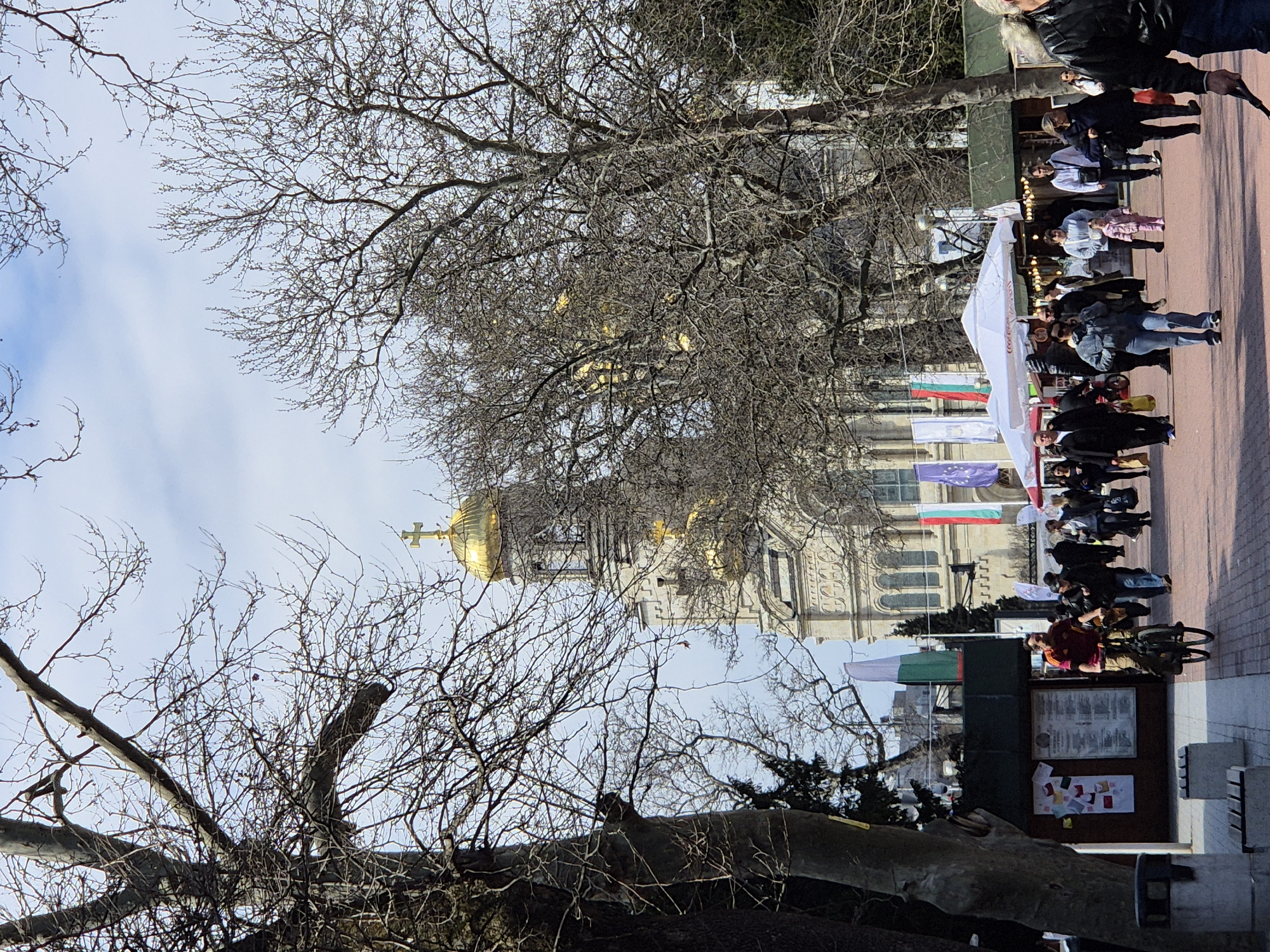 The image shows an outdoor scene in an urban setting. Dominating the center is a large building with a golden dome and cross visible through bare tree branches. In the foreground, people are walking along a sidewalk, some under a white tent structure. Flags of Bulgaria and the European Union are visible. A notice board stands near a tree trunk, and there are benches nearby.