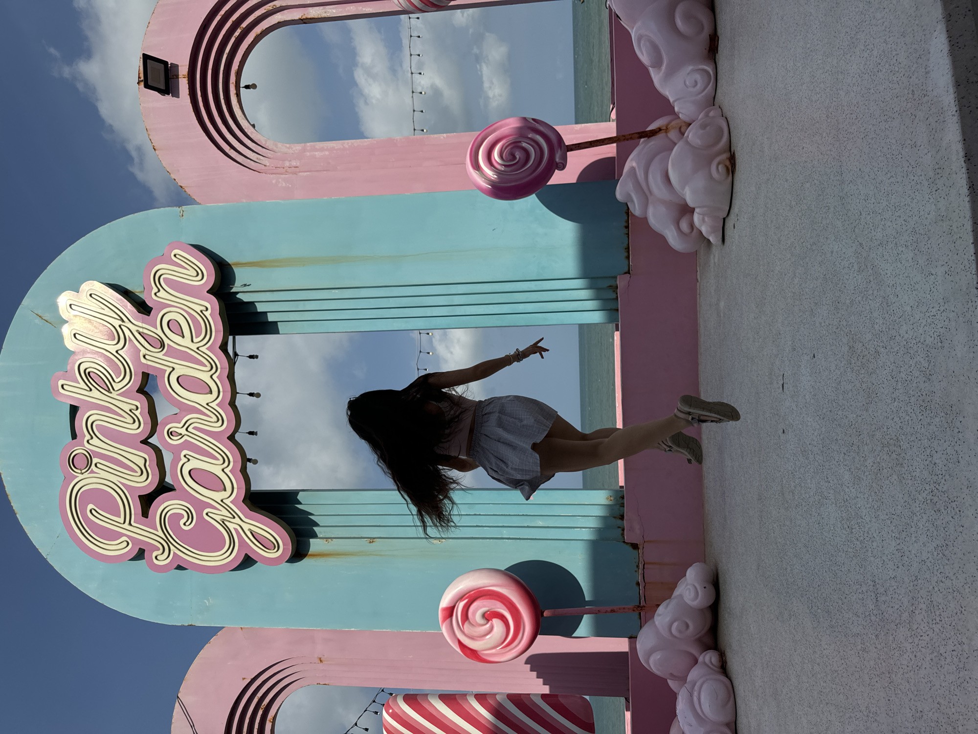 A woman with long, dark hair is captured mid-air, jumping in front of a vibrant pink and teal structure that reads "Pinky Garden" in cursive font. The structure features arched openings and is decorated with oversized candy-like props, including lollipops and striped candy canes. Pink, cloud-shaped sculptures are positioned at the base of the structure. In the background, the ocean meets the sky under a partly cloudy blue sky. The scene appears to be an outdoor attraction, possibly in a coastal area. The woman is wearing a light-colored top and a grey, pleated skirt.