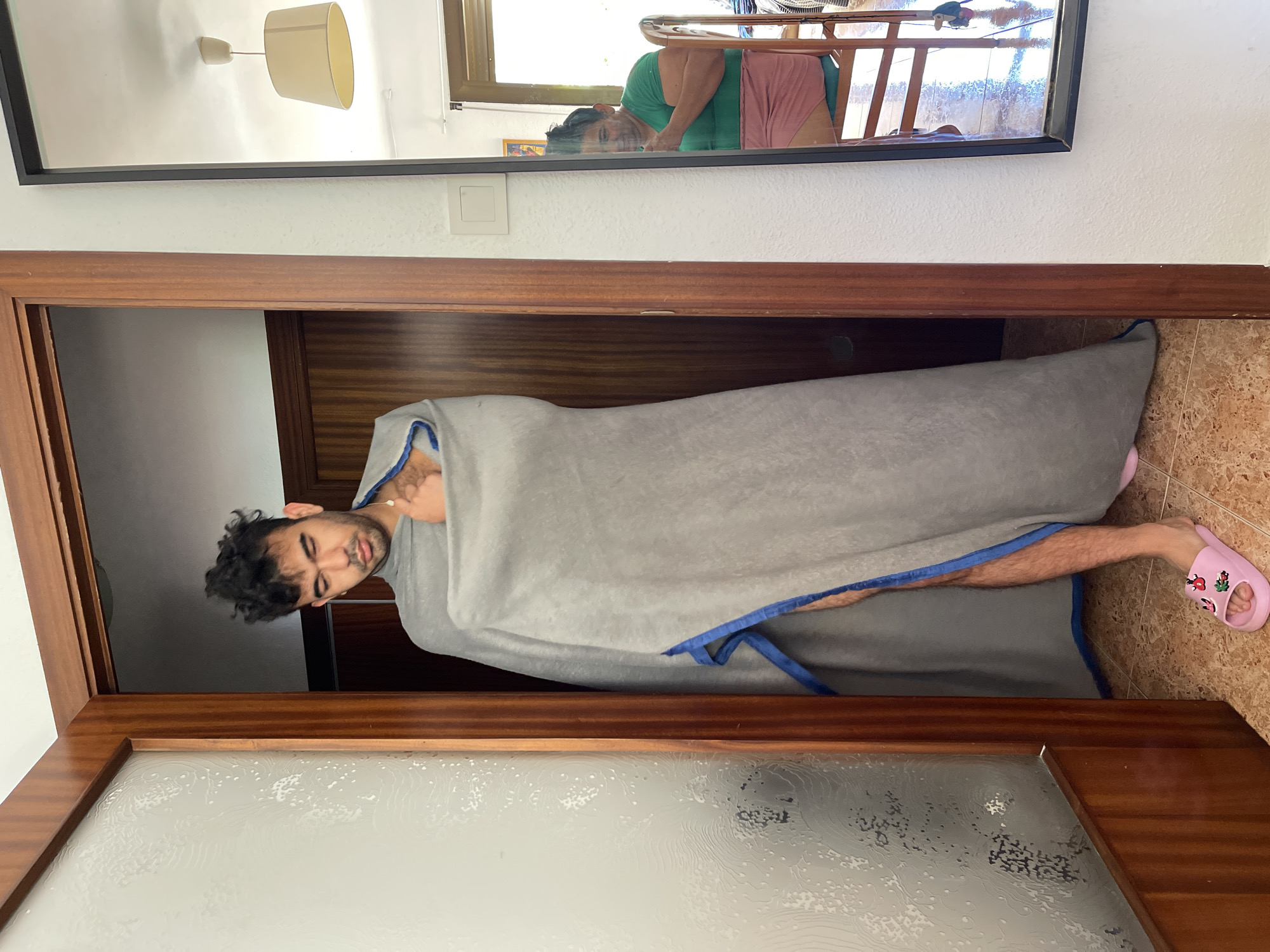 The image shows a man standing in a doorway, wrapped in a large gray blanket with blue trim. He is wearing pink slip-on sandals. His hair is dark and messy, and he has a pensive expression. To the right, a mirror reflects a woman sitting in a chair, wearing a green t-shirt and a pink skirt. The environment appears to be an interior, possibly a home, with wooden doorframes and a tiled floor. Based on the metadata, the location is Santa Coloma de Gramenet, Spain.