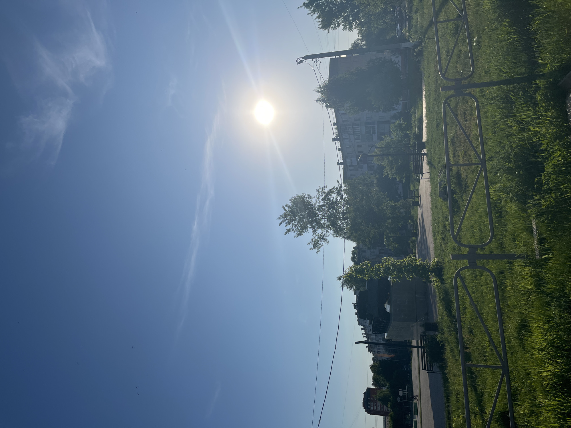 The image depicts a bright, sunny day in what appears to be a park or public space in Chaikovsky, Russia. A clear blue sky dominates the upper half of the image, with the sun positioned high, its rays causing a lens flare. The lower half shows a walkway with grass and a metal fence lining it. A few buildings and trees are visible in the background, along with power lines. The lighting conditions suggest it is daytime.