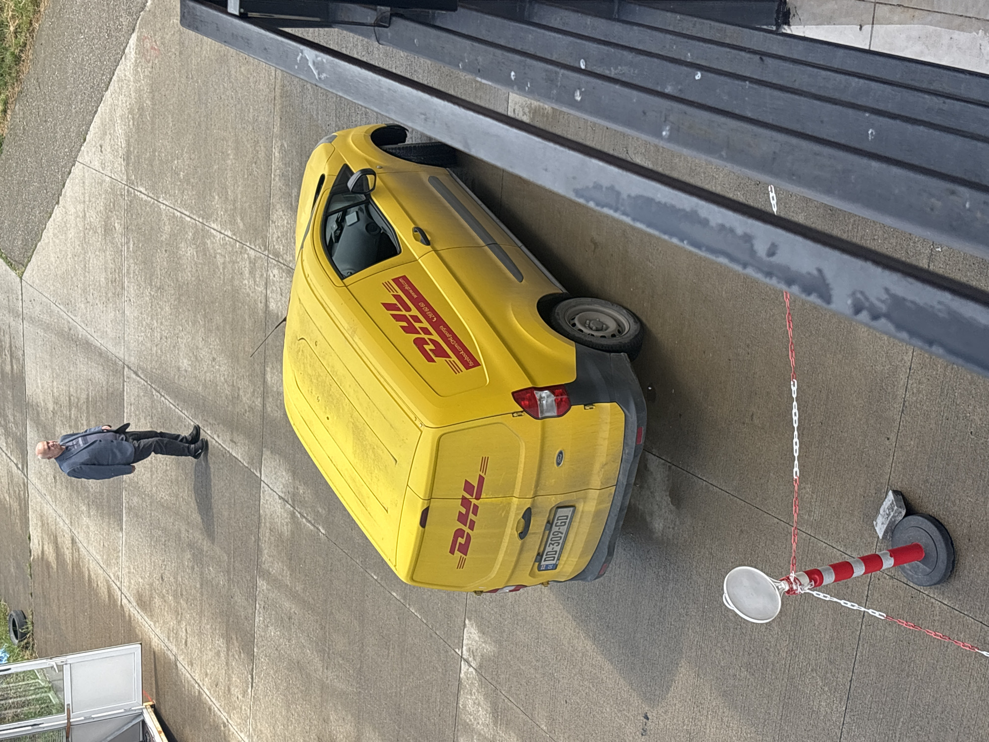 The image shows a yellow DHL delivery van parked on a concrete surface near a railing, a red and white striped bollard, and a man. The man, dressed in a dark suit, is walking away from the van. The license plate of the van is visible. The location cannot be determined based on the image.