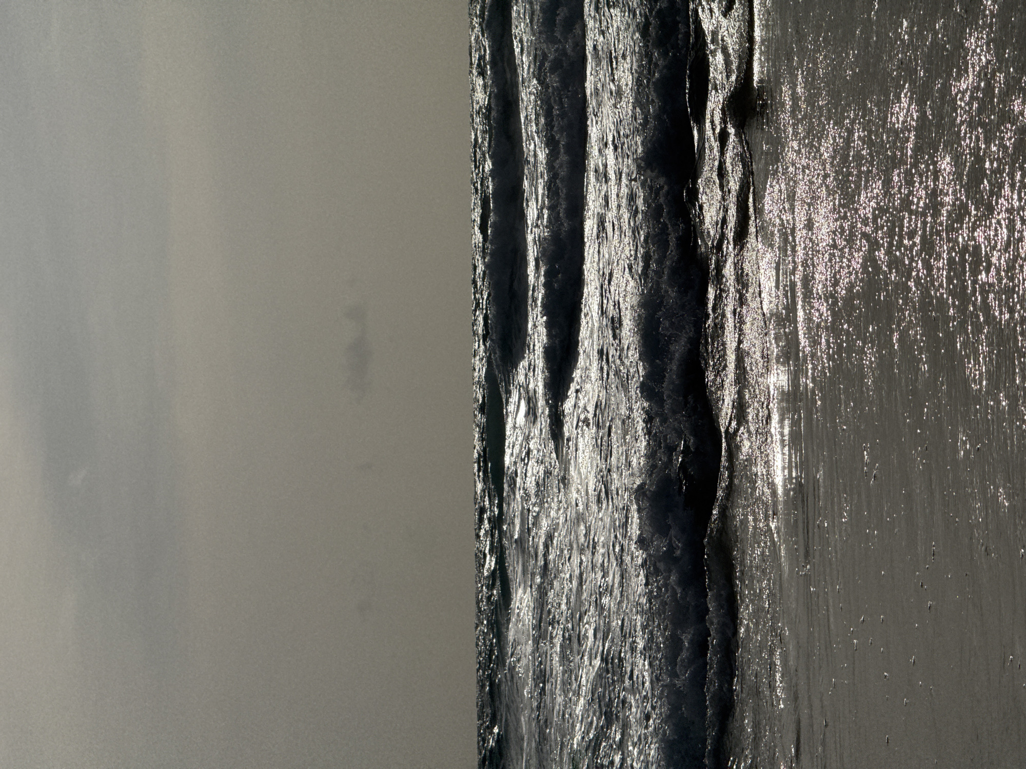 The image depicts a choppy sea with waves crashing against a sandy shore. The sky is overcast, casting a muted light over the scene. The water reflects the dim light, creating a shimmering effect on the surface of the waves and the wet sand. There are no people or man-made objects visible in the frame. The environment appears to be a natural beach scene. The image is a landscape shot taken at Karon Beach in Karon, Thailand.
