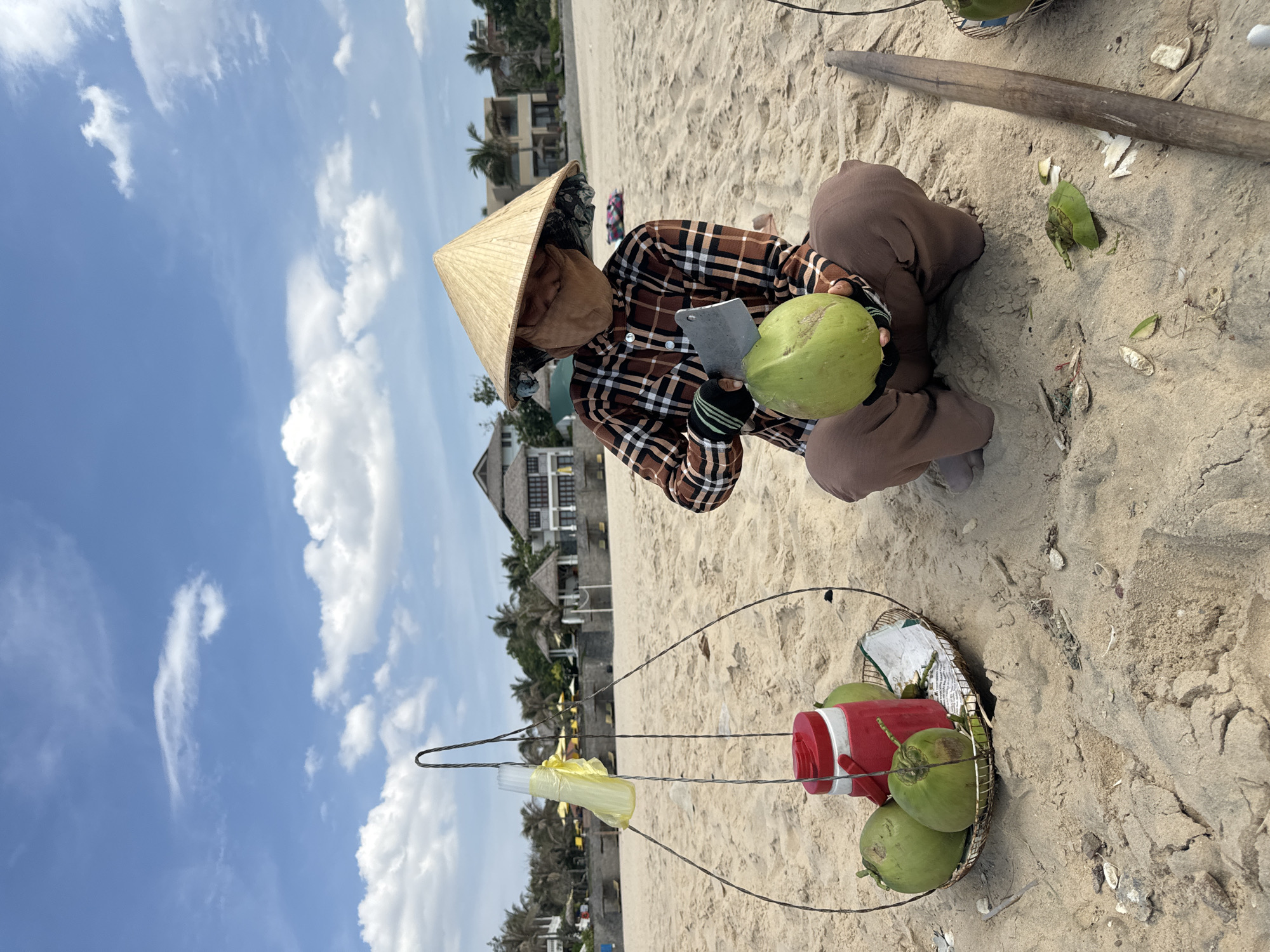 On a sandy beach in Phu Thuy Ward, Vietnam, a person wearing a conical Asian hat, a plaid shirt, and baggy brown pants squats on the sand, holding a green coconut in one hand and a cleaver in the other. A metal wire stand holds a basket containing additional green coconuts and a red container with a white lid. A bundle of yellow straws in a plastic bag hangs from the stand. The beach extends to the background with buildings and palm trees visible. The sky is blue with scattered white clouds. The scene is brightly lit by sunlight.