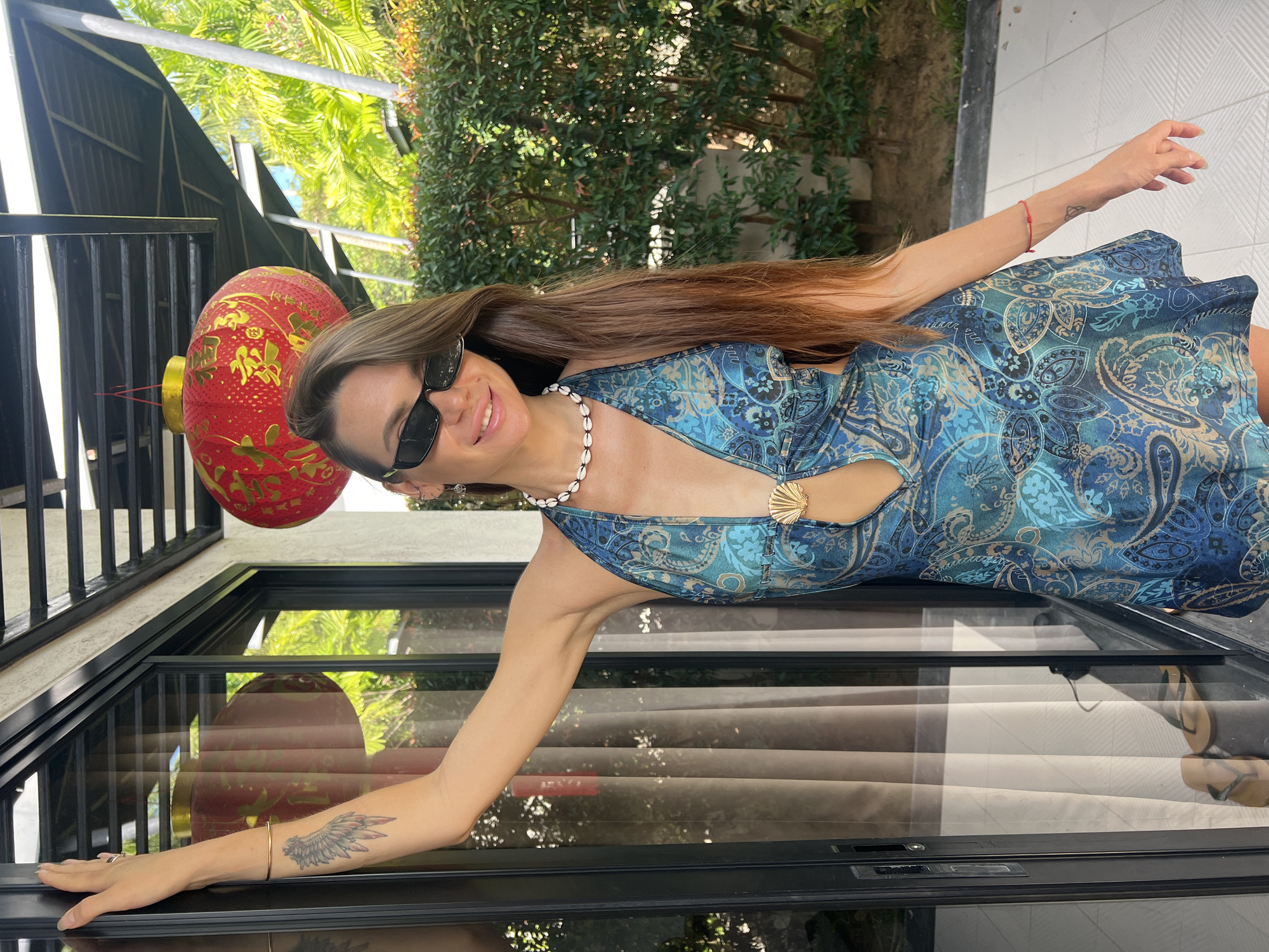 A woman with long brown hair, wearing sunglasses and a blue patterned halter dress with a seashell embellishment, stands leaning against a glass door. She has a tattoo on her left arm and a red bracelet on her right wrist. The background features lush green foliage, a red Chinese lantern hanging from a black awning, and a tiled floor with a pair of flip-flops visible through the glass door. The scene appears to be outdoors, possibly at a resort or residence in Ban Bang Rak, Thailand.