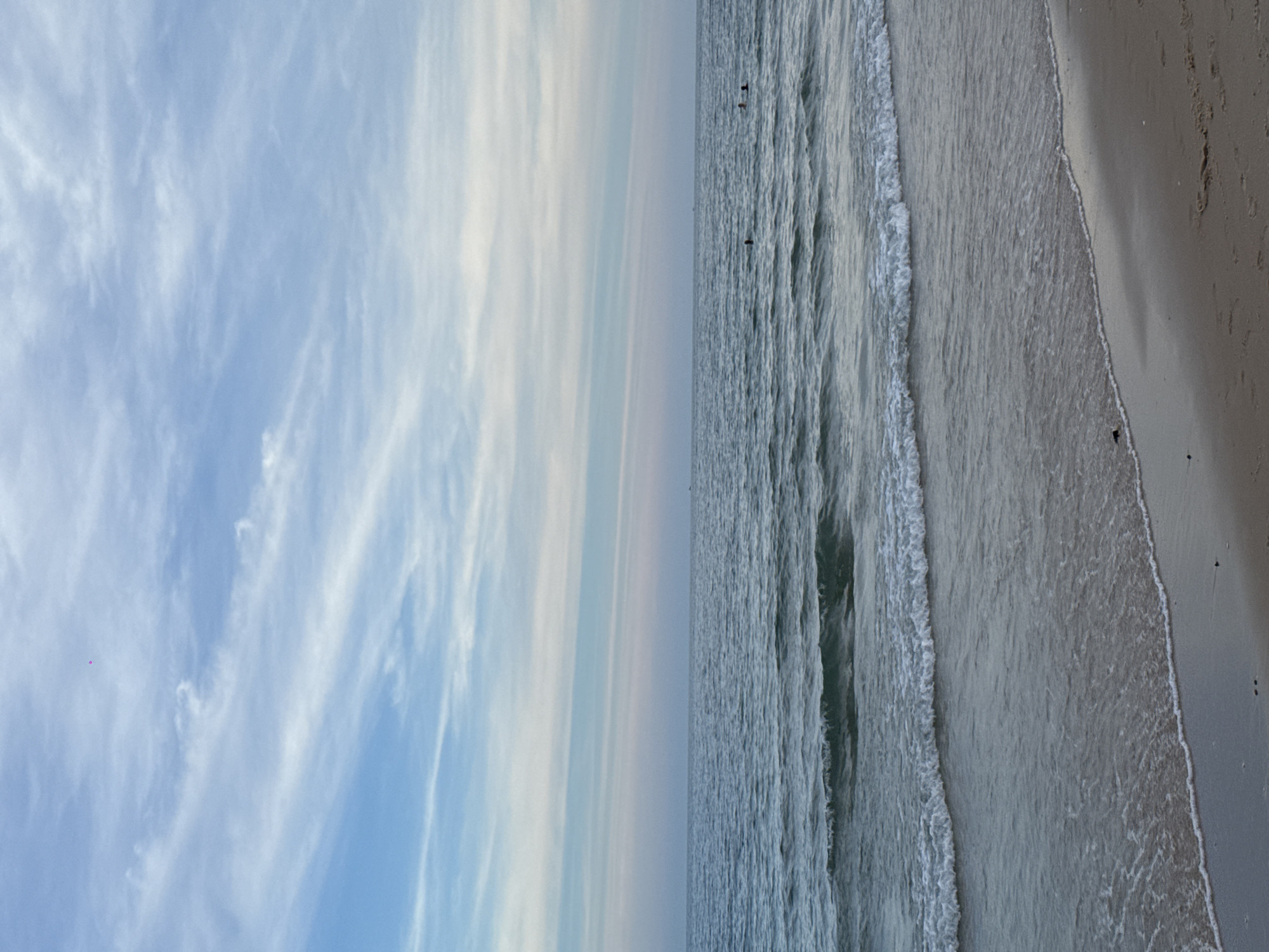 The image shows a serene beach scene at Phu Thuy Ward, Vietnam. The ocean is calm with gentle waves lapping at the sandy shore. The sky is a soft blue, with wispy white clouds stretching across the horizon. In the distance, a few small figures can be seen swimming in the water. The overall atmosphere is peaceful and tranquil, suggesting a perfect day for a beach outing. The sand is light brown and appears to be mostly clear, with a few small dark objects that could be shells or pebbles. The water is a muted blue-green, reflecting the sky above. A small pink object, possibly a kite, is visible high in the sky on the left side of the frame.