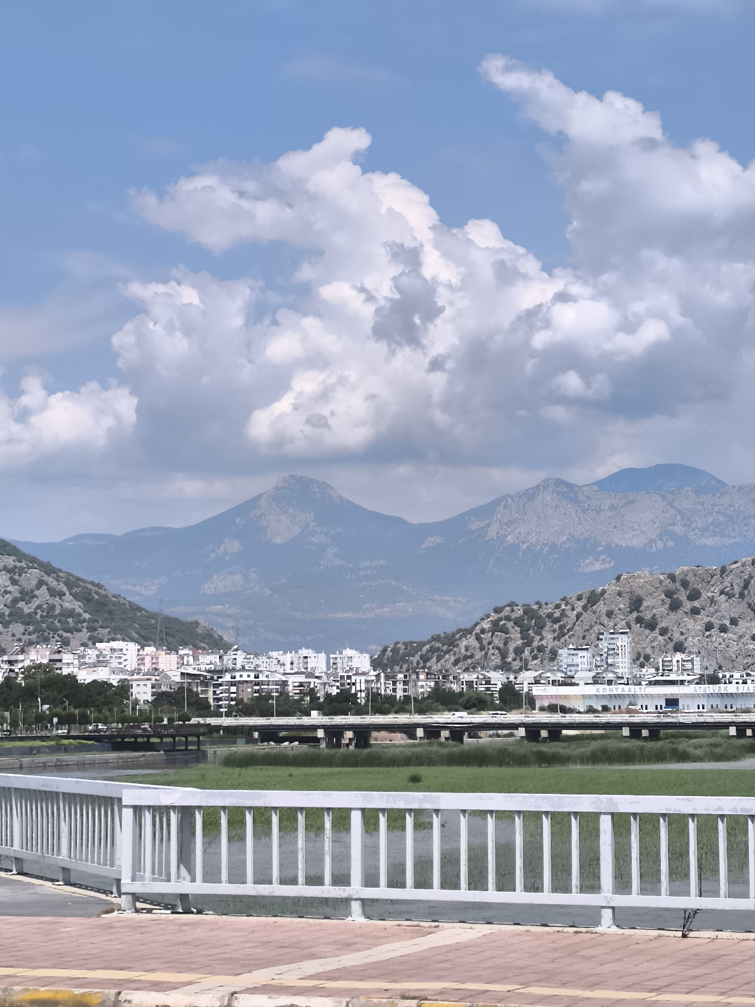 This image depicts a waterfront scene in Konyaaltı, Turkey, with a prominent white metal railing in the foreground. Beyond the railing, a body of water with a grassy embankment stretches into the middle ground. A bridge with multiple spans crosses the water, connecting the cityscape on the far bank. The cityscape is characterized by a dense collection of multi-story buildings, many appearing to be white or light-colored. In the background, a series of rugged, sparsely vegetated mountains rise against a sky filled with scattered white and gray clouds. The lighting suggests it is daytime, likely mid-morning or afternoon. No people or specific actions are discernible in the image.