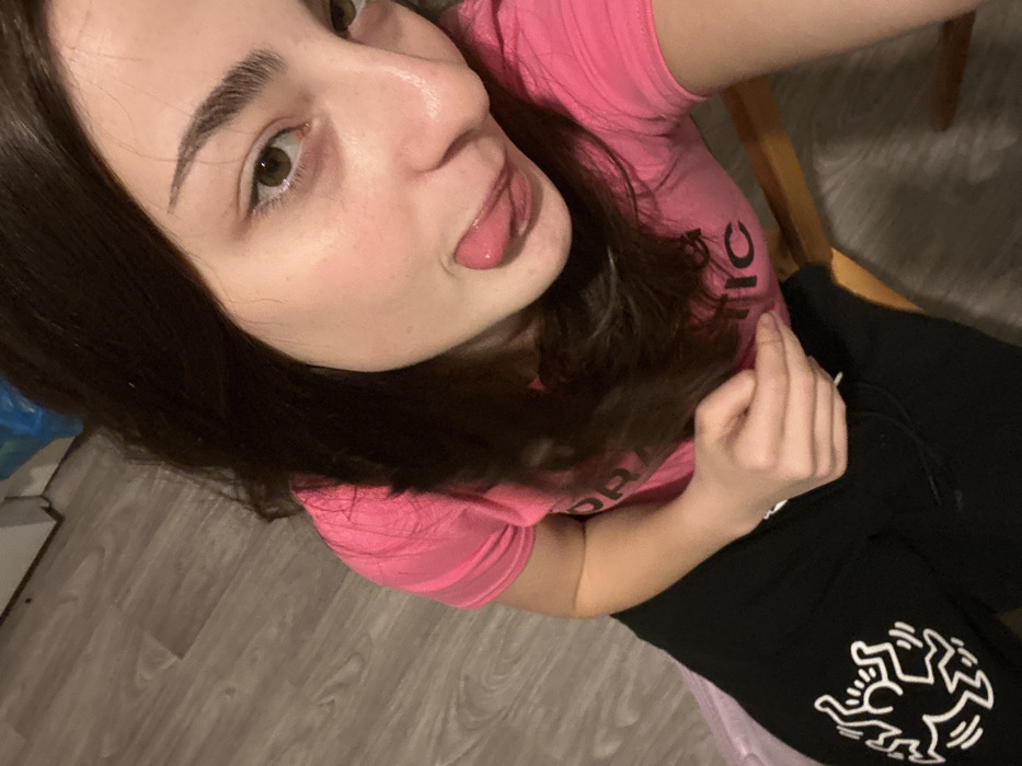 A young woman is seated, angled towards the camera in a selfie. She wears a pink t-shirt and black sweatpants featuring a white Keith Haring design. Her dark hair is loose, and she is sticking her tongue out slightly. Her expression is playful. The background shows a glimpse of light-colored flooring and a portion of a chair.