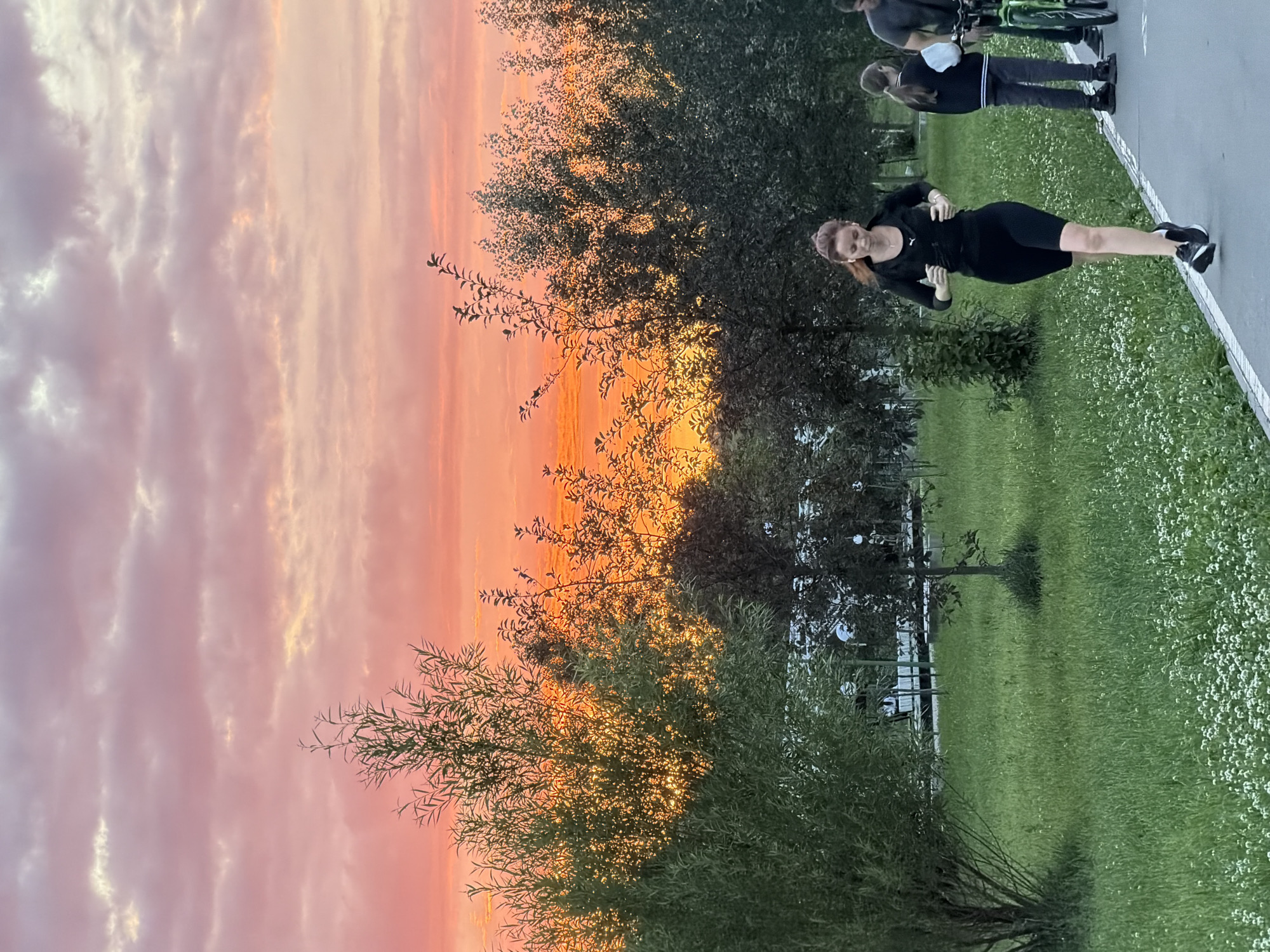 This media file shows a vibrant sunset with hues of orange, pink, and yellow clouds filling the sky. The foreground features a woman in a black athletic outfit, possibly exercising or posing for a photo, with her body angled towards the camera. She has red hair and is wearing black sneakers. In the background, there are trees with lush green foliage and a grassy area dotted with small white flowers. Other individuals are partially visible further in the background, one appearing to be standing near bicycles. The overall scene suggests an outdoor setting in a park or recreational area during sunset. The image is oriented vertically.