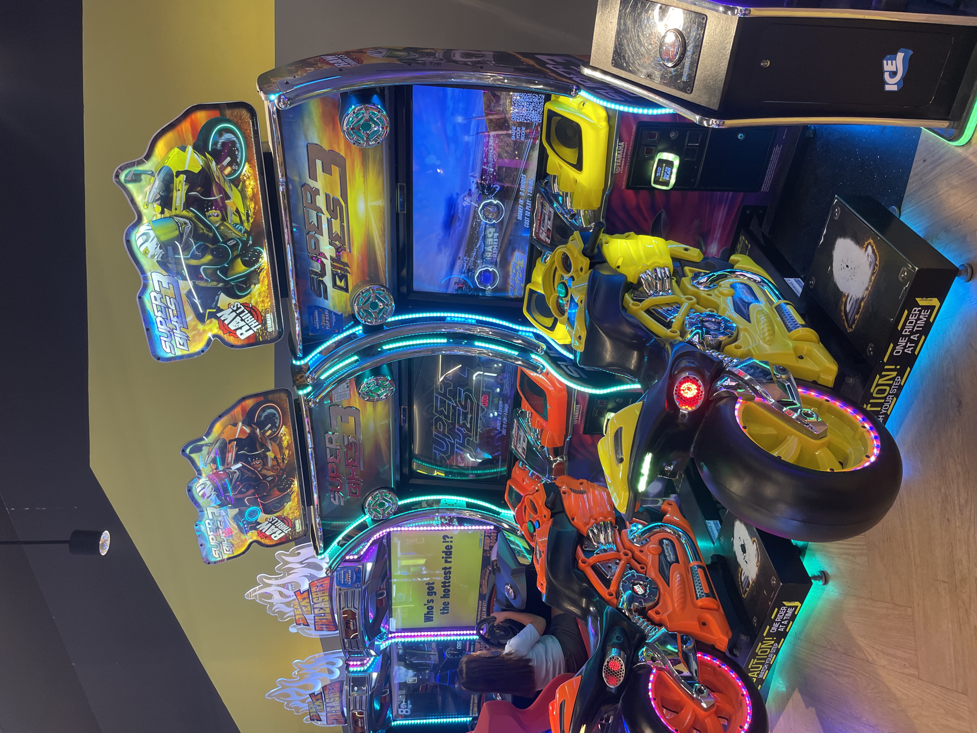 The image depicts a row of "Super Bikes 3" arcade racing game machines, commonly found in entertainment venues. One machine in the foreground features a bright yellow motorcycle seat, while another to its left has an orange motorcycle seat. A young woman, viewed from behind, is seated on the orange motorcycle, actively playing the game and holding the handlebars. The arcade machines have vibrant LED lighting, and the screens display gameplay graphics and game titles. The flooring appears to be light-colored wood. The background shows a wall with a yellow and black color scheme, and overhead lighting fixtures are visible. The overall environment suggests an indoor entertainment center. Based on the presence of "ICE" branding on a separate machine to the right, this arcade is likely located at the ICE (International Convention Exhibition) center in Manado, Indonesia, a popular venue for events and entertainment.
