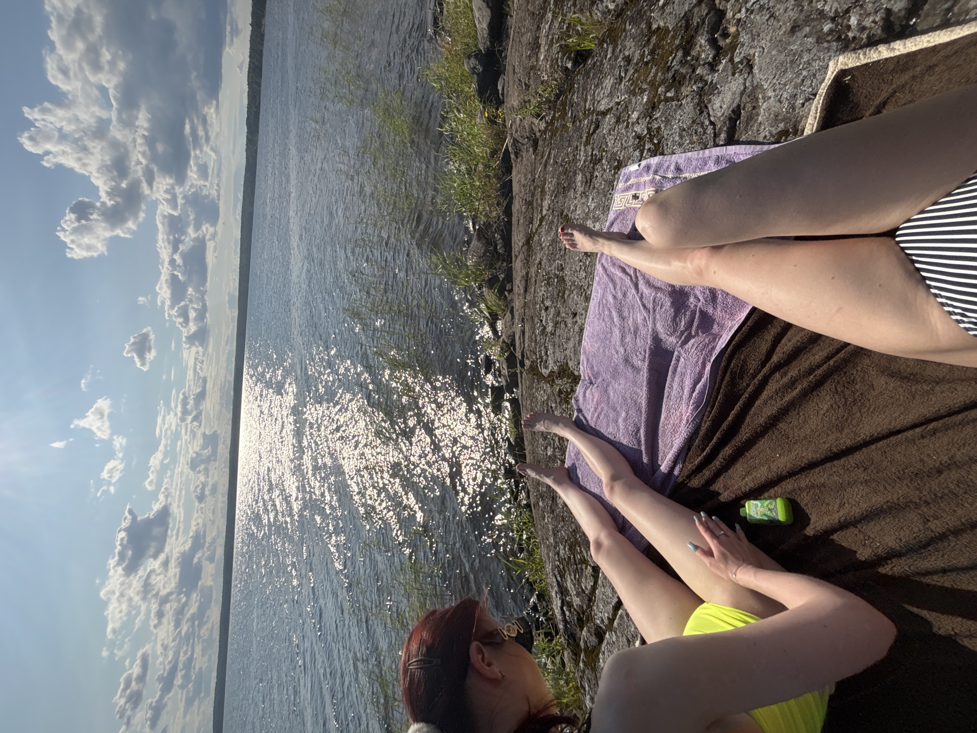 In an outdoor scene on a sunny day, two individuals are relaxing on a rocky shoreline next to a body of water. The person in the foreground, viewed from the waist down, wears a black and white striped bikini bottom and lies on a brown towel. Their legs are extended, with their right foot resting on a purple towel. To the left, a second person, viewed from the side and slightly from behind, is seen from the upper torso down to their legs. They have red hair and are wearing a bright yellow bikini top. Their legs are also extended, and their hands are resting on their lap. A small green bottle, possibly sunscreen, is placed on the brown towel between them. The background features a vast expanse of water reflecting the sunlight, with a distant treeline and a bright sky filled with scattered white clouds. The rocky terrain suggests a natural, outdoor setting.