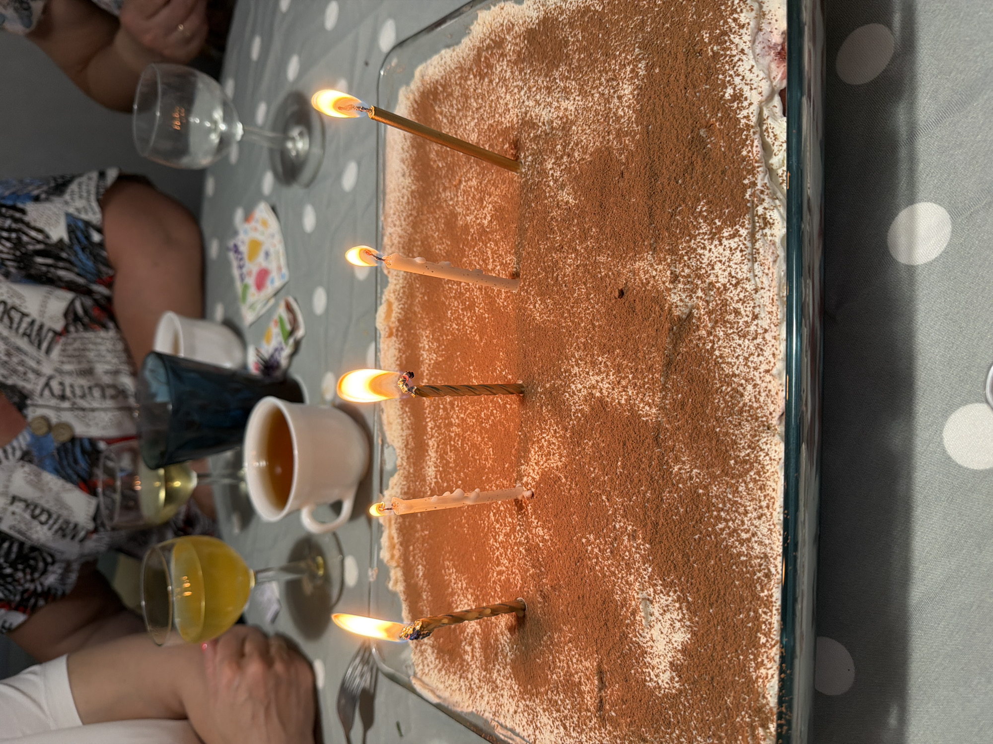 The image depicts a rectangular tiramisu cake in a glass dish with five lit candles, likely part of a birthday celebration. The cake is dusted with cocoa powder and appears to be served at a table. Around the cake are various items: a glass of yellow liquid, a clear wine glass, a white mug containing a light-colored beverage, a dark blue glass vase, and playing cards. At least two people are visible, one wearing a garment with a newspaper print and another wearing a white shirt. The table is covered with a gray tablecloth adorned with white polka dots. The location, Votkinsk, Russia, cannot be confirmed from visual information alone.