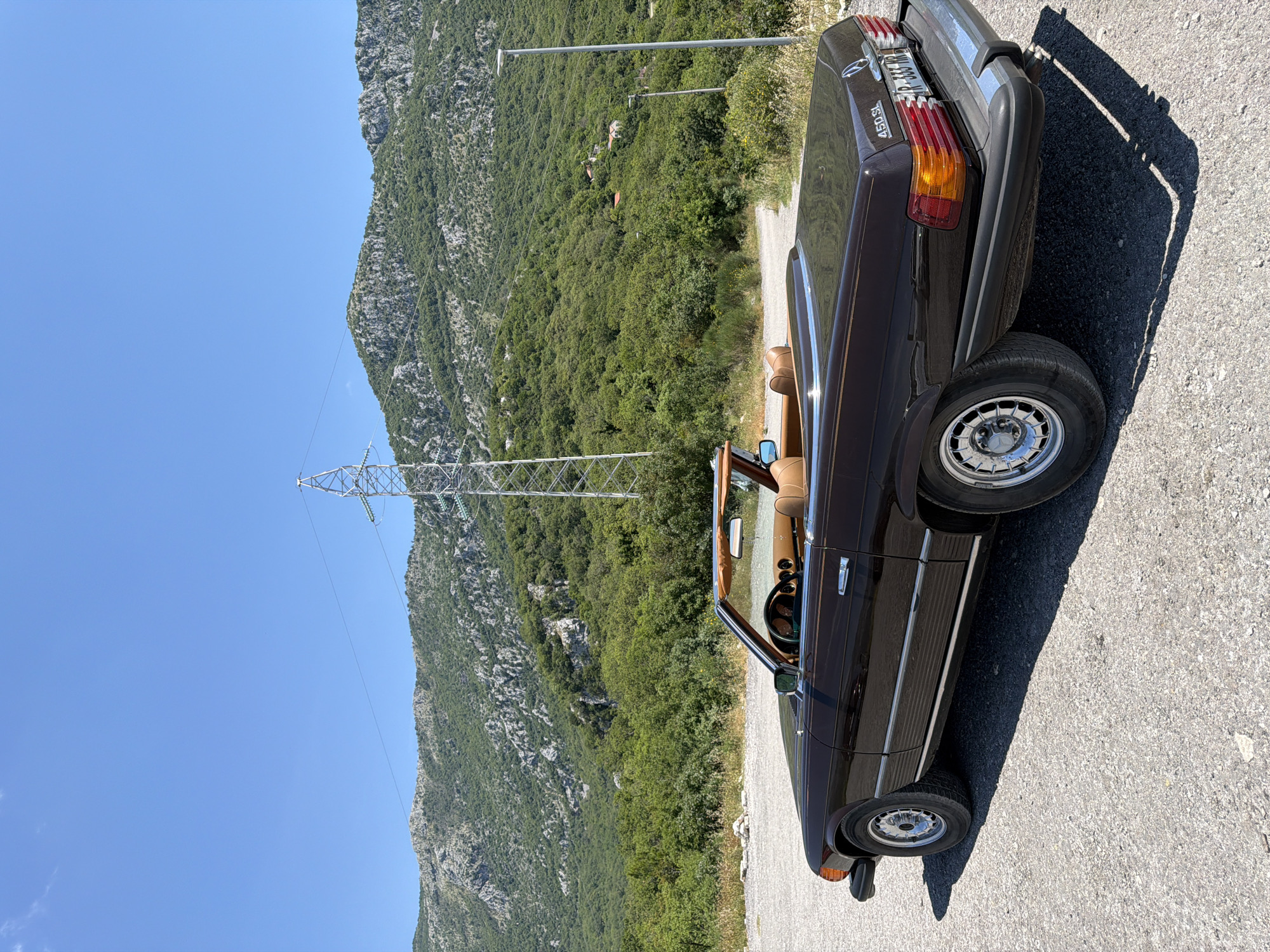 The image shows a brown 1970s Mercedes-Benz 450 SL convertible parked on a gravel road. The car is facing right, with its top down, revealing tan leather interior. In the background, there is a lush green mountain landscape under a clear blue sky. A tall, metal power line tower stands prominently to the right of the car, with wires stretching across the frame. The area appears to be rural, with sparse vegetation and a few scattered rocks on the ground. The sunlight casts a sharp shadow of the car on the road. The license plate on the car reads "R 333 HR". The location is identified as being in Sveti Stefan, Montenegro. There are no people or any discernable actions taking place in the image.