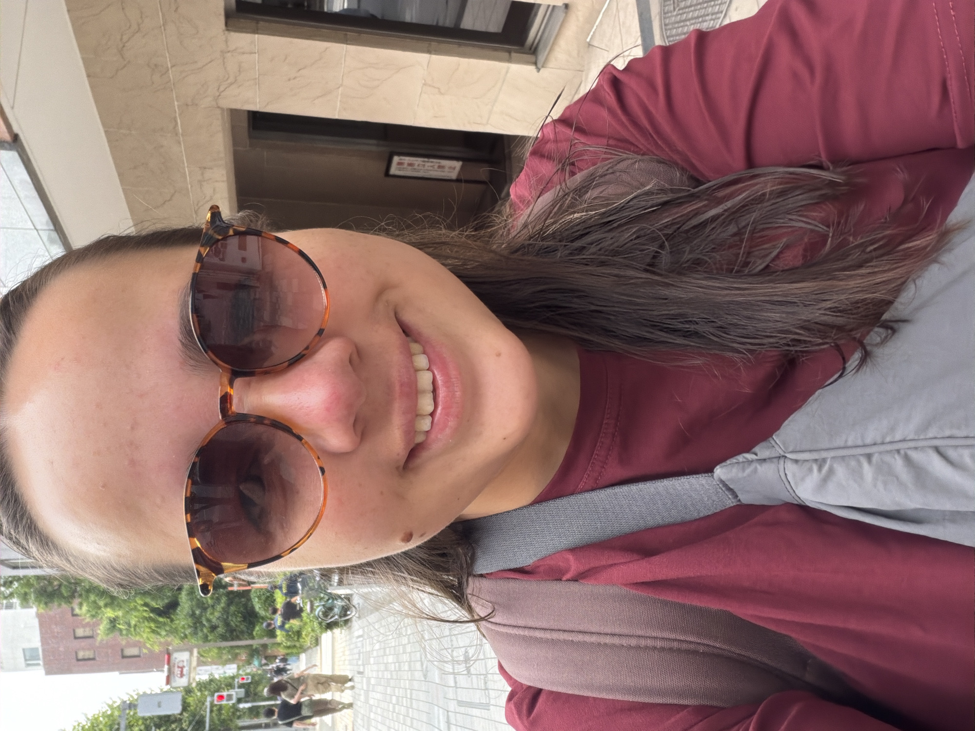 A selfie-style, medium shot shows a young woman with brown hair and fair skin, wearing sunglasses with tortoise shell frames and a maroon t-shirt. A gray cross-body bag with tan straps is visible over her shoulder and chest. She is outdoors in what appears to be a city, with a sidewalk and buildings in the background. In the background, two people are walking on the sidewalk, and a bicycle is parked near a building. The environment suggests an urban setting in Chuo, Japan, likely during daylight hours.