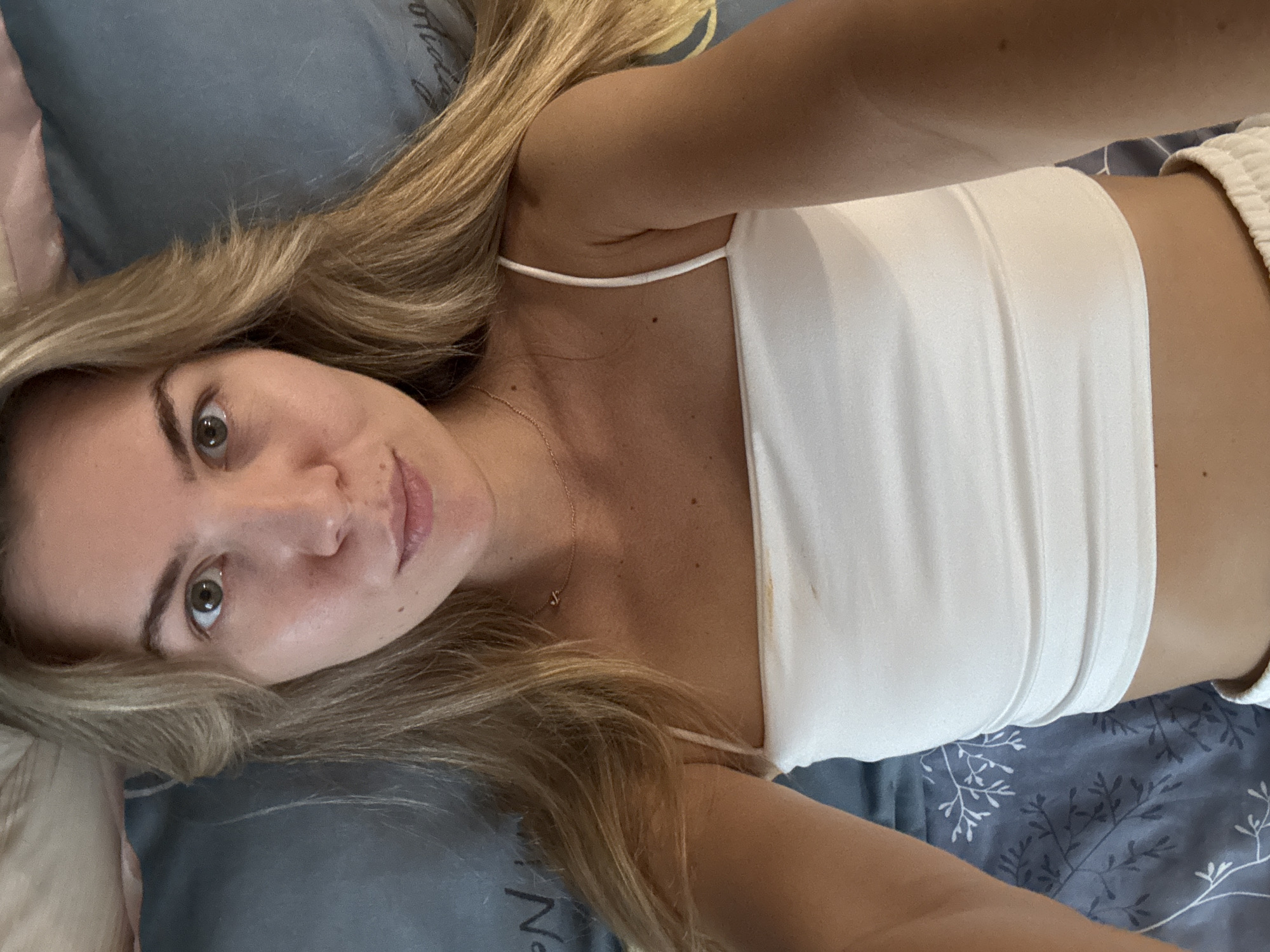 A blonde woman lies on a blue and gray patterned bed, looking at the camera. She has light skin, brown eyes, and her blonde hair is parted to the left. She is wearing a white tank top and white shorts. Her arms are outstretched, suggesting she is taking a selfie. The background is a bed with a patterned duvet. There is some text written in what appears to be cursive on the duvet.