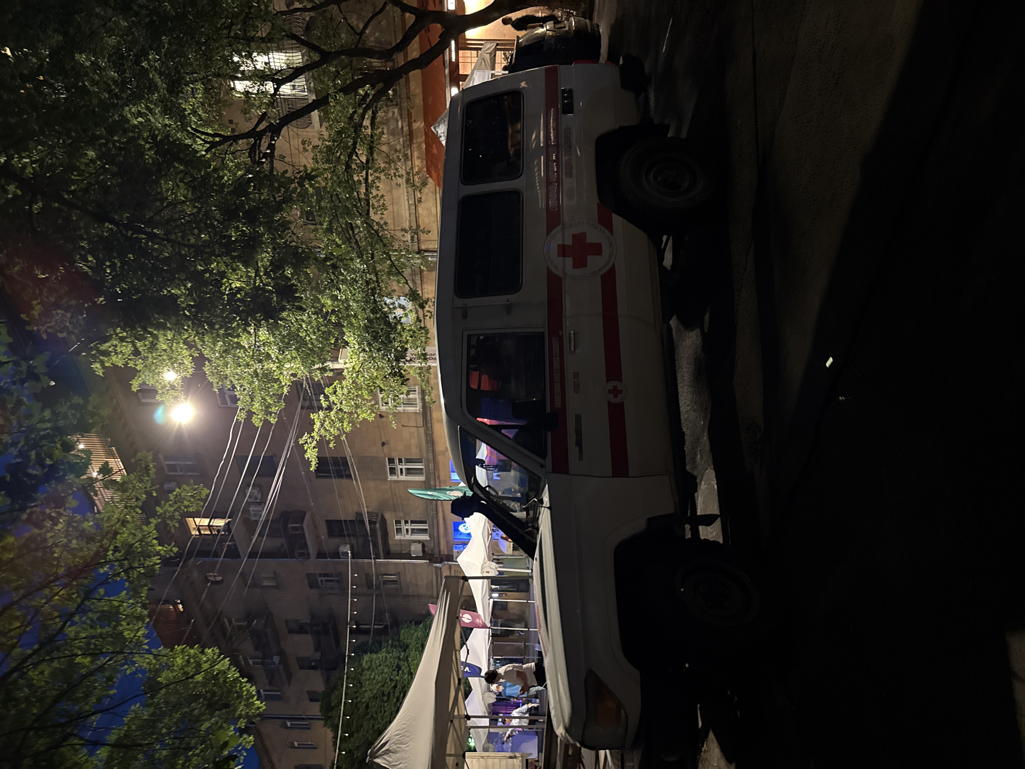 The image captures a nighttime street scene in Yerevan, Armenia. A white utility vehicle, clearly identifiable as belonging to the Armenian Red Cross, is prominently parked on the right side of the frame. The vehicle features a distinctive red stripe running along its side and the iconic red cross emblem. Text in Armenian on the vehicle reads "ՀԱՅԱՍՏԱՆՅԱՆ ԿԱՐՄԻՐ ԽԱՉԻ ԸՆԿԵՐՈՒԹՅՈՒՆ" (Armenian Red Cross Society) and "ՀԱՅԱՍՏԱՆՅԱՆ ԿԱՐՄԻՐ ԽԱՉ" (Armenian Red Cross). Additionally, "AMBULANCE" and "ՇՏԱՊ ՕԳՆՈՒԹՅՈՒՆ" (Emergency Aid) are visible on its front door, confirming its role.
In the mid-ground, several white canopy tents are set up, under which people are gathered, suggesting an outdoor event or temporary station. One person wearing a light blue shirt is clearly visible within one of the tents.
The setting is an urban environment at night, illuminated by a bright streetlight on the left, as well as lights emanating from the tents and multiple windows of the buildings in the background. Lush green trees with dense foliage line the street, partially obscuring some of the buildings. Numerous power lines are visible crisscrossing the upper part of the scene against the dark night sky. The atmosphere appears calm, indicative of an organized evening event or community presence by the Armenian Red Cross.