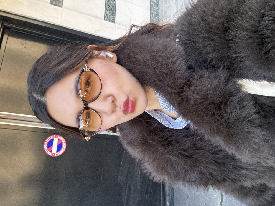 A frontal portrait captures an adult female in a selfie pose on a street in Paris, France. She is wearing dark, oval-shaped sunglasses with amber-tinted lenses, which reflect surrounding architecture. Her lips are pursed and covered in red lipstick. A white earbud is inserted into her right ear. She is dressed in a textured, dark brown fur-like coat over a light blue and white striped collared shirt. In the immediate background, to her left, is a dark, possibly metallic, surface. Affixed to this surface is a red, white, and blue circular regulatory sign with French text indicating "EMPLACEMENT RÉSERVÉ" (Reserved Spot) and "STATIONNEMENT INTERDIT" (No Parking). To her right, a building facade composed of light-colored panels with dark, possibly green-veined, horizontal accents is visible. The scene is illuminated by natural daylight.