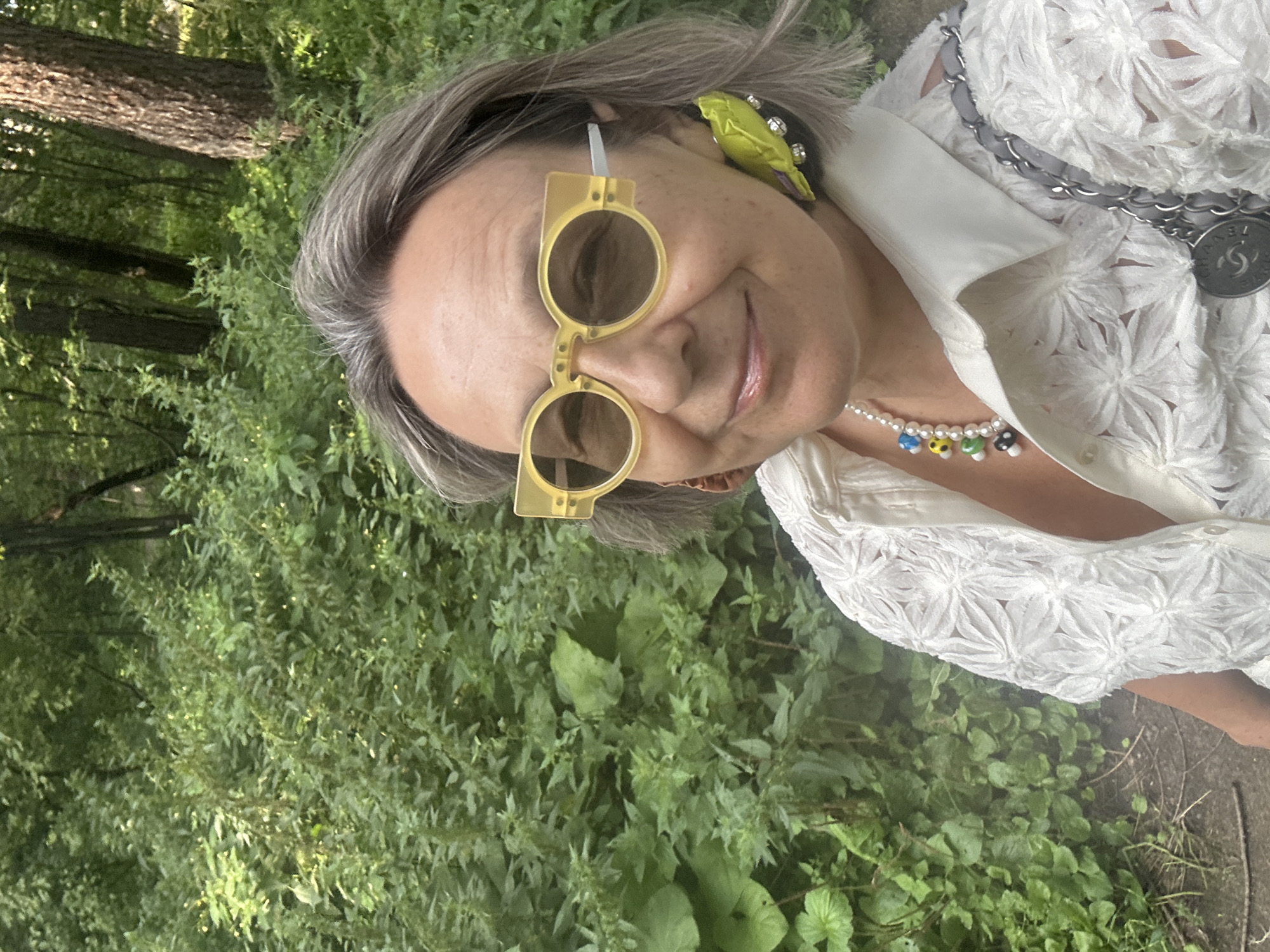 A woman with gray hair and light-colored skin is taking a selfie outdoors in a wooded area. She is wearing large, yellow, rectangular-framed sunglasses and a white blouse with an intricate floral cutout pattern. She has a pearl necklace with colorful mushroom-shaped beads around her neck and large, bright green statement earrings. A gray chain bag with a Chanel logo charm is slung over her right shoulder. The background is filled with lush green foliage and trees.
