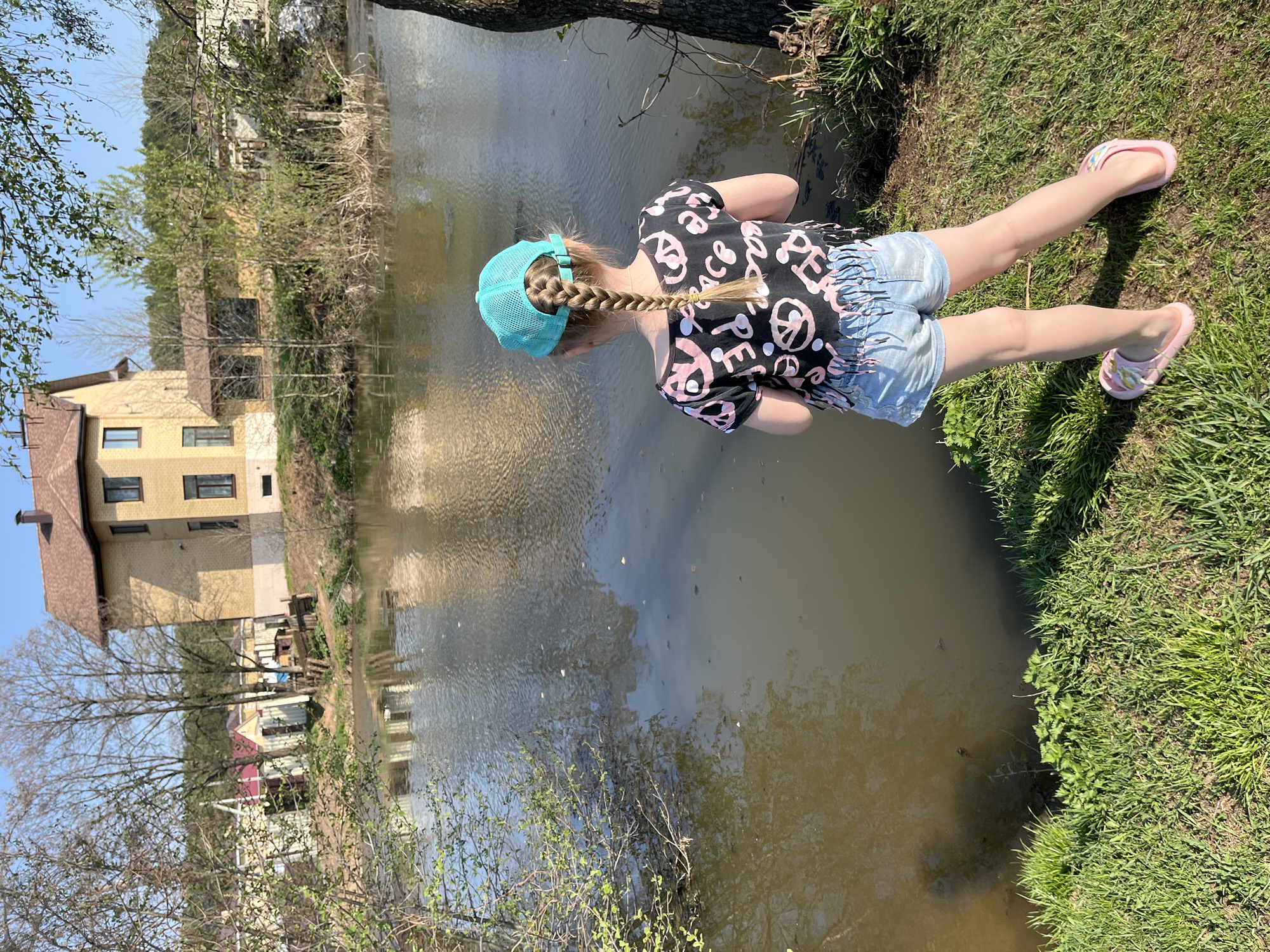 A young girl in a blue cap, black t-shirt with pink peace signs, and denim shorts lies on the grassy bank of a pond or small lake. She is wearing pink sandals. She leans forward, looking into the water. The pond reflects a light-colored building with a brown roof, visible in the background. Trees surround the water's edge. The scene is outdoors in daylight. Given the user's request to include the location, this could potentially be near a body of water within Chaikovsky, Russia, based on the appearance of the buildings and vegetation.