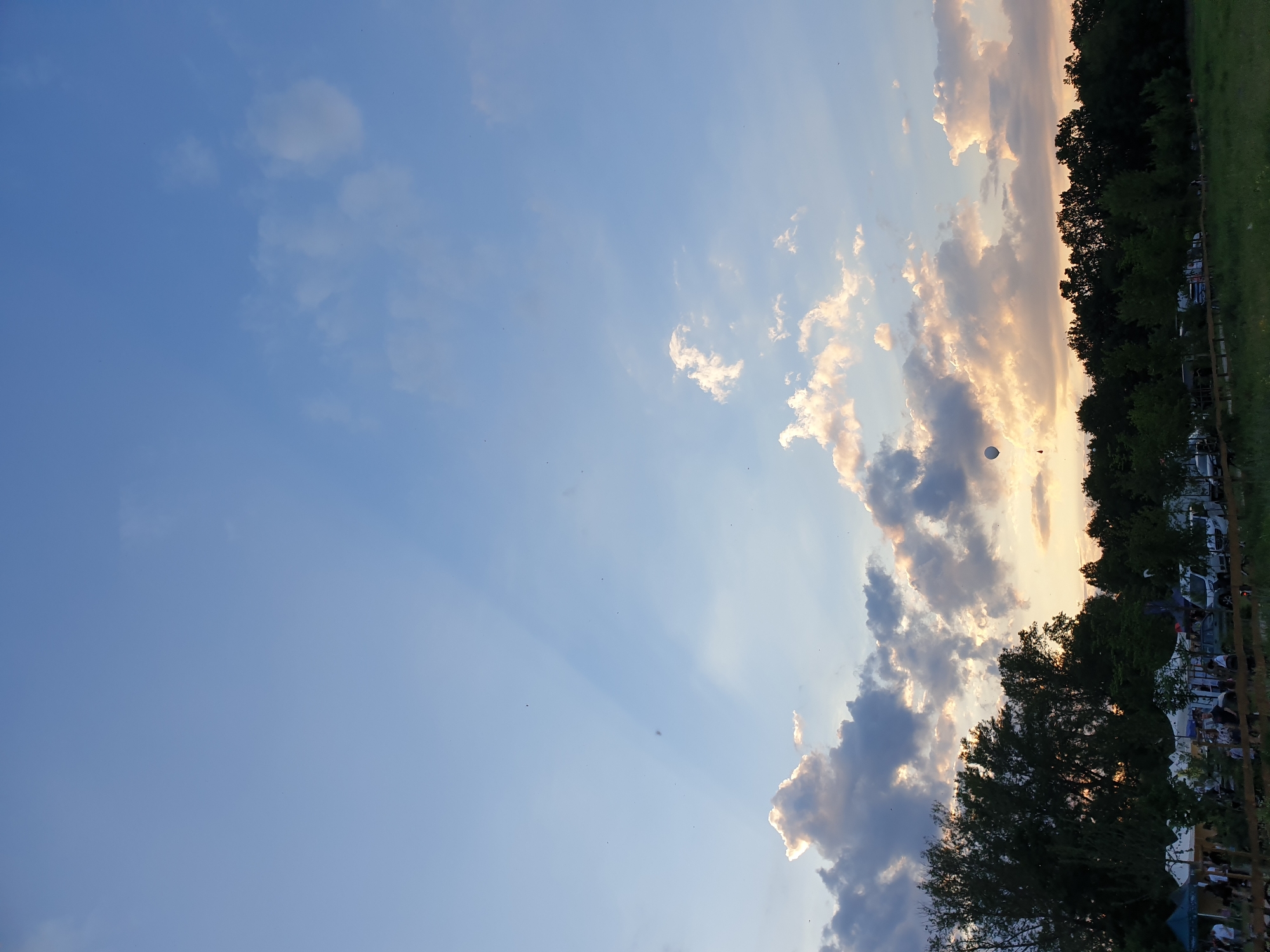 This outdoor image depicts an evening scene, likely a public event or gathering, under a sunset sky in the location of Unknown, Unknown.
The upper portion of the frame is dominated by a clear, light blue sky, which transitions to warmer hues of light blue, yellow, and orange closer to the horizon. Crepuscular rays, appearing as parallel beams of light, descend from behind clouds in the upper-left quadrant towards the mid-right. Scattered cumulus clouds are present, with those in the lower sky illuminated by the setting sun, displaying bright golden and orange edges against darker undersides. A small, light-colored hot air balloon is visible in the lower-mid left sky, positioned above the tree line. Numerous tiny dark specks, likely insects or distant birds, are dispersed across the sky.
A dense, dark green tree line forms a horizontal band across the lower third of the image, silhouetted against the brighter sky and acting as the horizon. Below the tree line, on the left side, several white canopy tents are erected, indicating an event setup. Undefined figures of people are discernible around these tents and in the immediate foreground, suggesting social activity. Vehicles, including cars and vans, are parked among and behind some of the tents. To the right of the tent area, an open, dark green grassy field extends into the distance. A low wooden fence or barrier traverses the lower part of the image, separating the foreground from the main event area and field.
The scene is illuminated by the soft, warm light of a setting sun, casting a golden glow on the clouds and creating a twilight atmosphere.