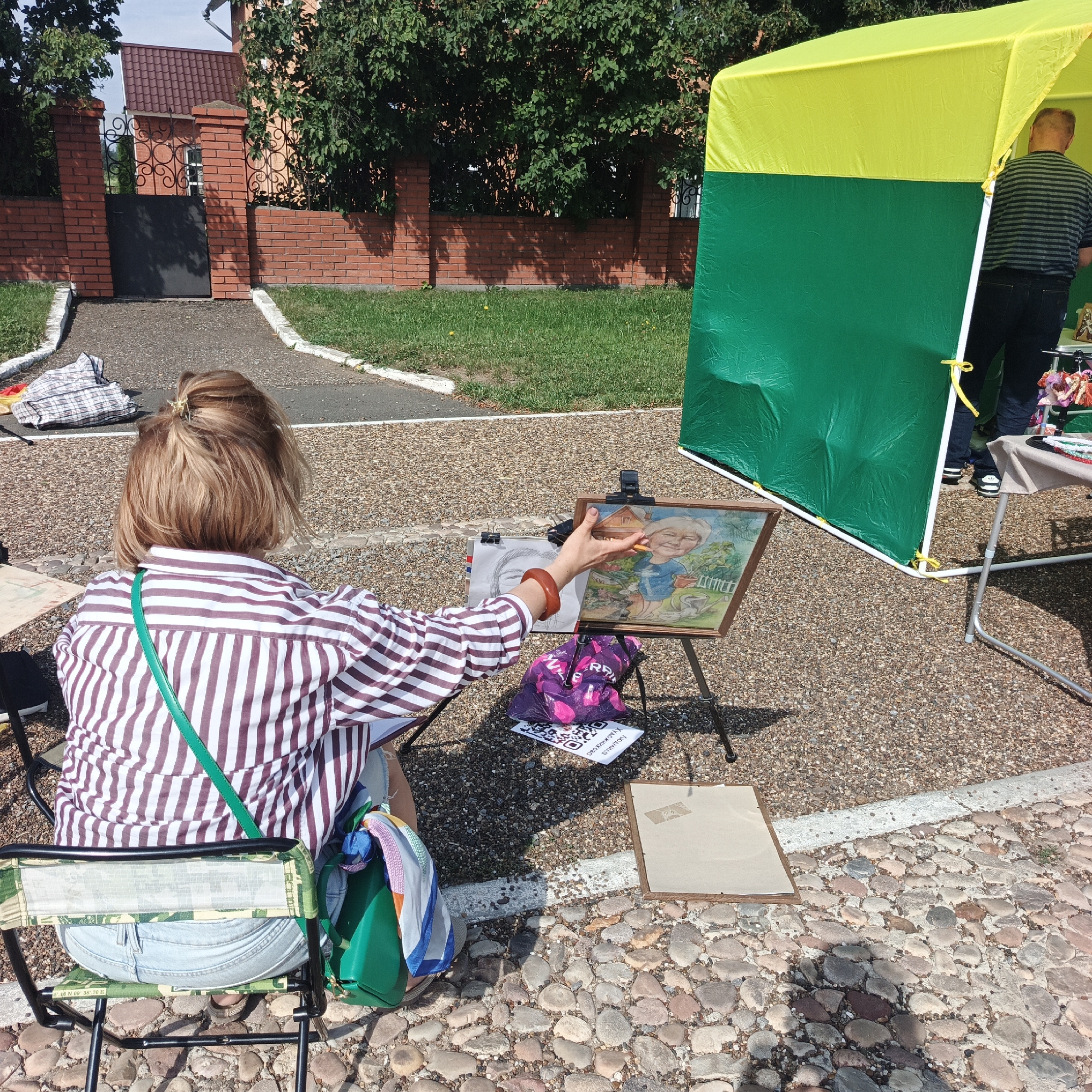 In Nikolo-Beryozovka, Russia, a woman sits on a foldable chair, facing away from the camera, as she draws on an easel. She is wearing a striped shirt and denim shorts, with a green strap draped across her body. The easel displays a framed drawing of an elderly person. To the right, a yellow and green tent is set up, partially obscuring a man in a striped shirt and jeans. The ground is covered with small stones, and in the background, a brick fence and trees are visible. The scene suggests an outdoor art event or market.