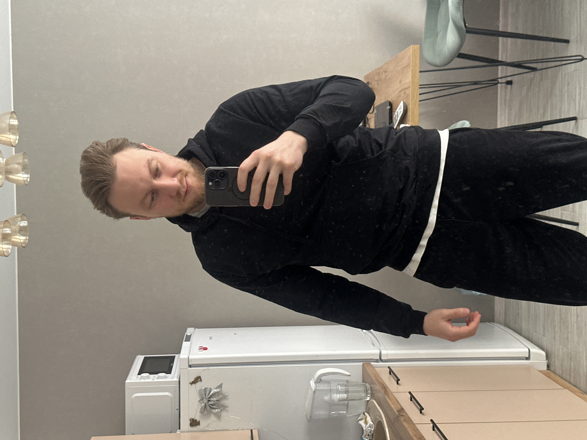 A man in a black tracksuit is taking a selfie in a kitchen. The kitchen includes a white refrigerator with decorations, a washing machine, beige cabinets, a wooden table, and a blue chair. The location is Sochi, Russia.