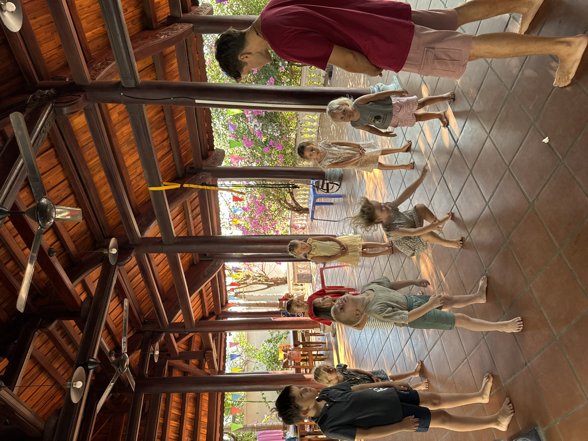 The image shows a group of people, primarily children, standing in a row on a tiled floor, possibly engaged in an activity or game, likely a line dance. An adult is visible at the front of the line and the back of the line, seemingly guiding or participating. The location is under a wooden-beamed structure with fans, suggesting an outdoor covered area. Decorative elements, such as plants and colorful decorations, are visible in the background. Based on the architecture and decorations, this is most likely occurring in Mũi Né, Vietnam, due to the prevalence of similar architectural styles in the area.