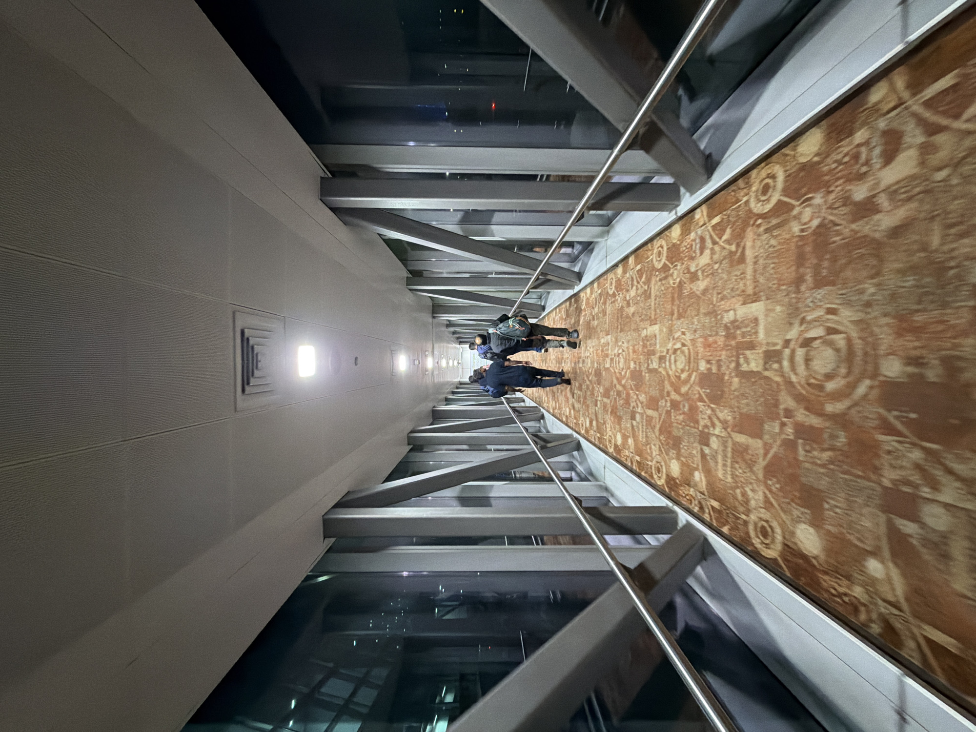 The image shows an interior view of a jet bridge at an airport in New Delhi, India. The jet bridge has a long, symmetrical corridor with a patterned carpet on the floor. The walls are made of glass panels supported by metal beams, offering views of the airport tarmac and lights at night. Several people are walking away from the camera down the jet bridge. The lighting is artificial and recessed into the ceiling.
