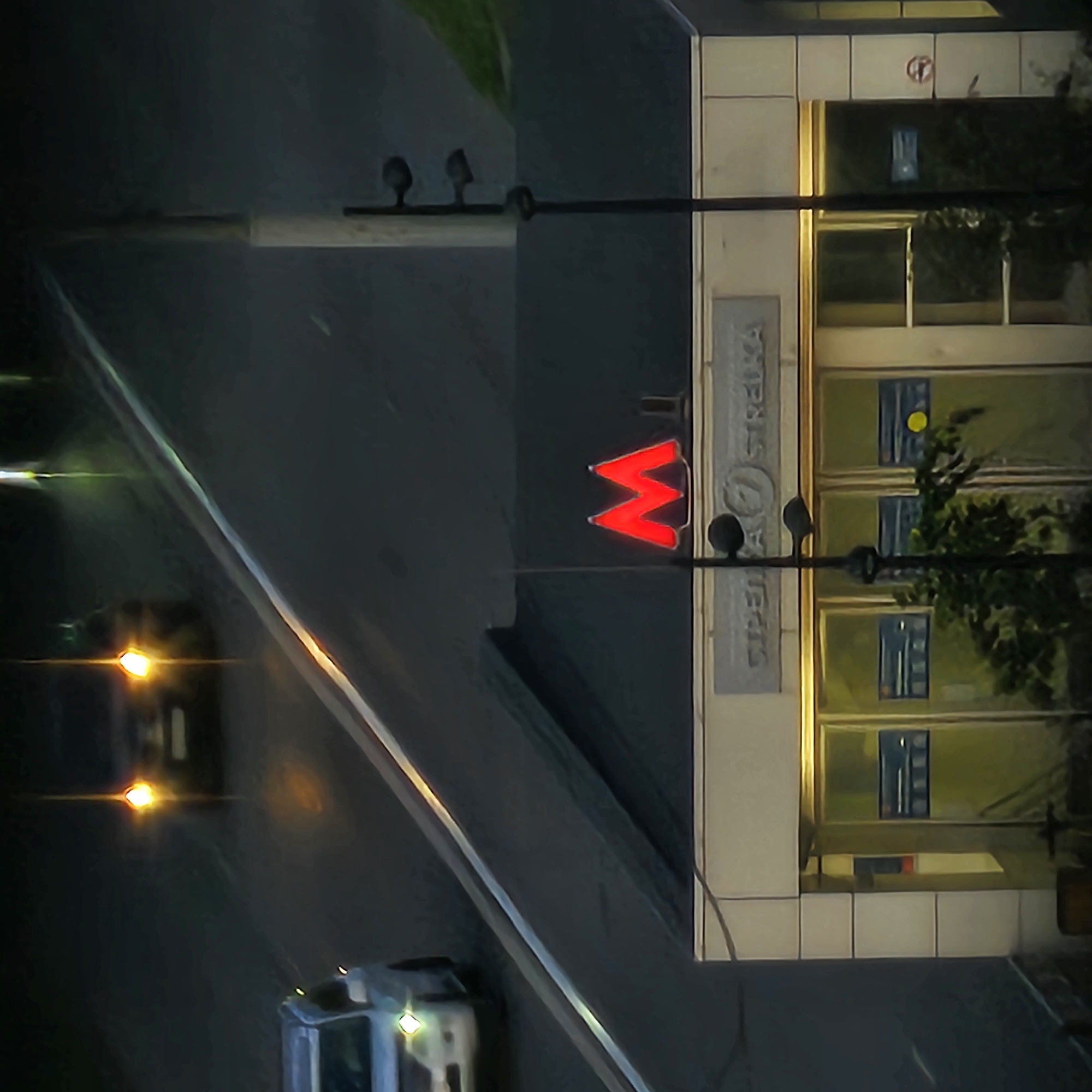 This is a high-angle, rotated shot of a street scene at night, likely in Nizhny Novgorod, Russia. The primary focus is a modern building with large windows and a prominent red "M" logo, indicating a metro station entrance. The building has a light-colored facade with tiled sections. Vertical text is visible on the building's facade, appearing to read "СТРЕЛЬНА" and "СУДЕБНАЯ".
In the foreground, a dark road with a light-colored stripe runs diagonally across the image. Two cars are visible on the road. One car, further away and to the left, has its headlights on, casting a bright glow. A second car, closer and to the right, is partially visible, appearing to be a light-colored utility vehicle with its lights on.
The scene suggests a city environment at night, possibly after dark, given the artificial lighting from the cars and the metro station. The weather is not discernibly indicated. The overall mood is urban and somewhat stark due to the low light conditions and the high-angle perspective.