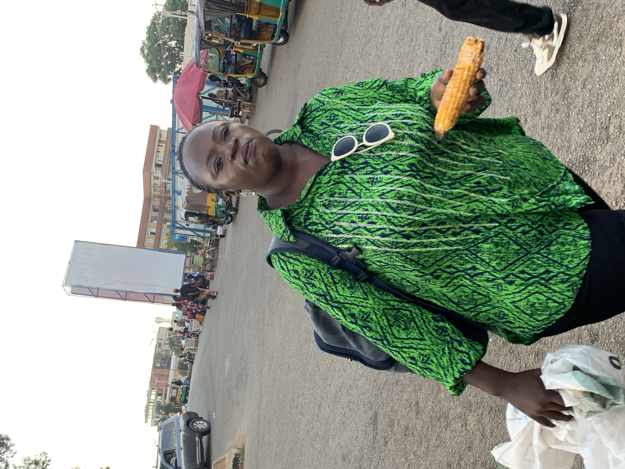 An individual stands in a dusty, outdoor urban environment, likely a market or roadside area in Jos, Nigeria. The individual, a woman with dark skin and braided hair, is facing the camera with a neutral expression. She is wearing a vibrant green shirt with a black and white patterned design and a black backpack slung over her left shoulder. A pair of white-rimmed sunglasses is perched on the collar of her shirt. In her right hand, she holds a cooked ear of corn. Her left arm is bent as she clutches a white plastic bag filled with items.
The background reveals a street scene with vehicles, including a dark-colored SUV on the left and several colorful auto-rickshaws parked along the right side of the road. Other individuals are visible in the distance, some appearing to be vendors or shoppers. A large, white billboard stands prominently in the background. The ground is a rough, grey asphalt surface. The overall lighting suggests it is daytime, possibly late afternoon.