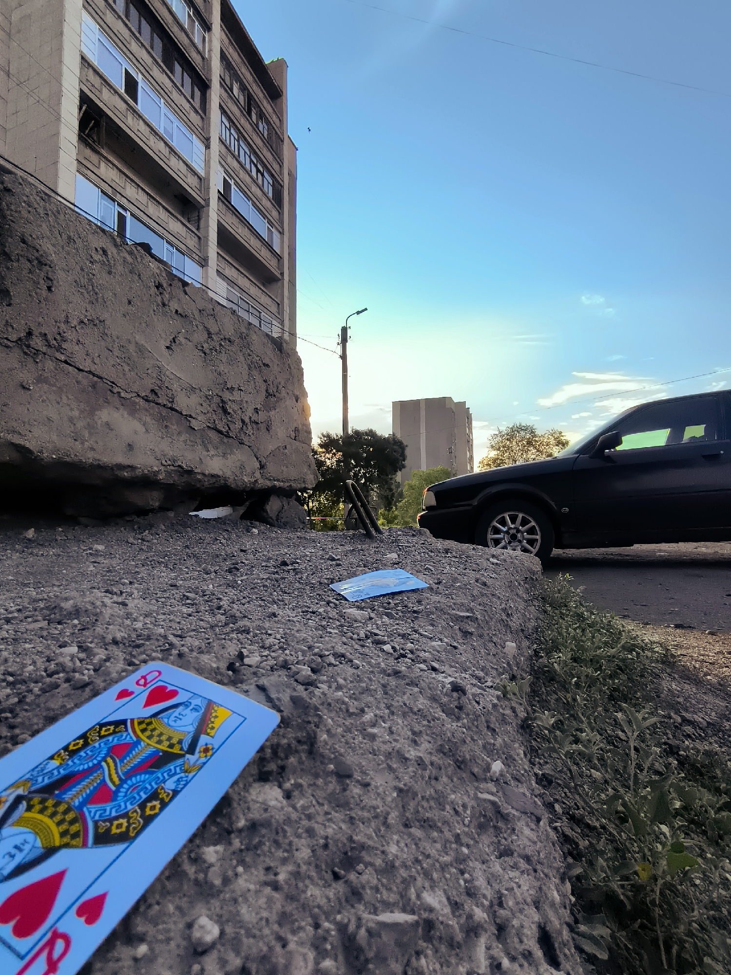 The image depicts an outdoor scene in Qarağandı city administration, Kazakhstan. In the foreground, a playing card, the Queen of Hearts, lies face up on a rough, gravelly surface. To the left, a large, weathered concrete structure dominates the lower portion of the frame, showing signs of cracking. Behind this structure, a multi-story building with a light-colored facade and rows of windows is visible. The sky above is a clear, pale blue, with the sun setting and casting a warm glow. A black car is parked on a paved road to the right, with some green vegetation growing along the edge of the road. Further in the background, another tall building and trees can be seen. A utility pole with wires is also present.