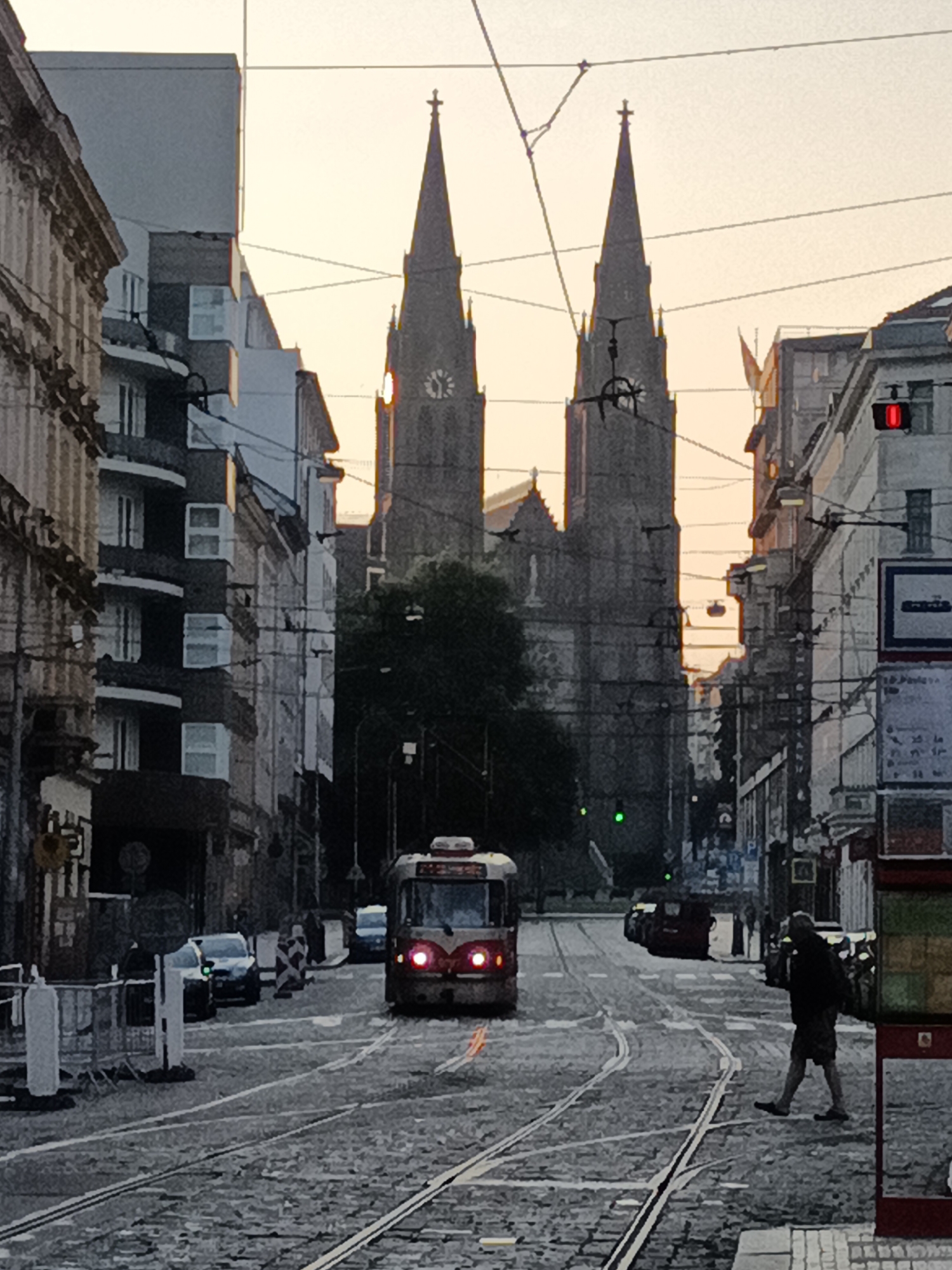 The image depicts a street scene in Prague, Czechia, at what appears to be dusk or dawn, with the sky a soft orange. In the background, two prominent church spires, characteristic of Prague's architecture, dominate the skyline. A red tram is visible in the middle of the cobblestone street, its headlights illuminated. Other cars and pedestrians are also present, contributing to the urban atmosphere. The street is lined with buildings, some of which have balconies and visible windows. Overhead, tram wires crisscross the sky, indicating the presence of public transportation. The overall impression is that of a bustling city transitioning between day and night.
