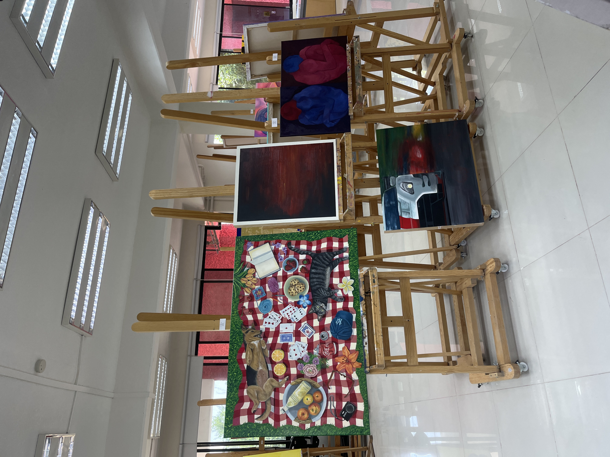 The image shows a collection of paintings on wooden easels in a brightly lit room with fluorescent lights on the ceiling. The floor is tiled with white squares. The paintings depict various subjects: a picnic scene, an abstract image of figures, a dark red abstract, and an image that includes a robotic figure. The paintings are arranged vertically on multiple easels. The room appears to be a studio or gallery space, possibly located in San Andrés Cholula, Mexico, given the prompt's instructions. The scene includes paintings on easels, and the focus is on the display of art. There are no people visible in the image.