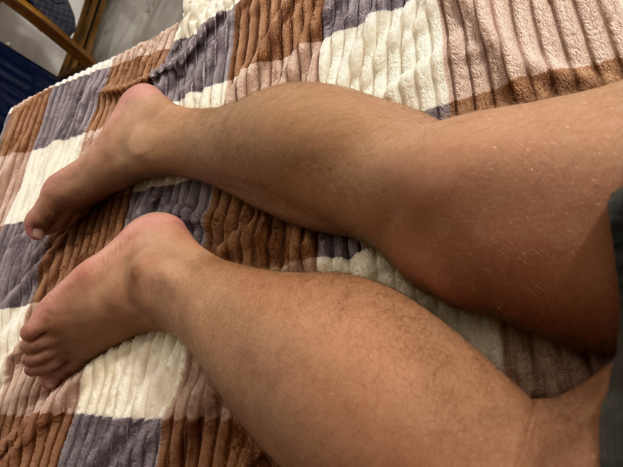The media file shows a close-up view of a person's legs and feet resting on a textured blanket. The blanket has a pattern of alternating bands of brown, beige, and white. The skin on the legs is tanned and appears to have fine hairs. The toes of the feet are visible, with one foot showing the top and the other showing the side. The lighting suggests an indoor setting, possibly in the evening or with artificial light, as there are no strong shadows or natural light cues. The overall scene is calm and relaxed, with no specific activity or event taking place. No text is visible in the image. The provided location context, Kadewatta, Sri Lanka, does not offer any specific visual cues within the image itself to confirm or deny it.