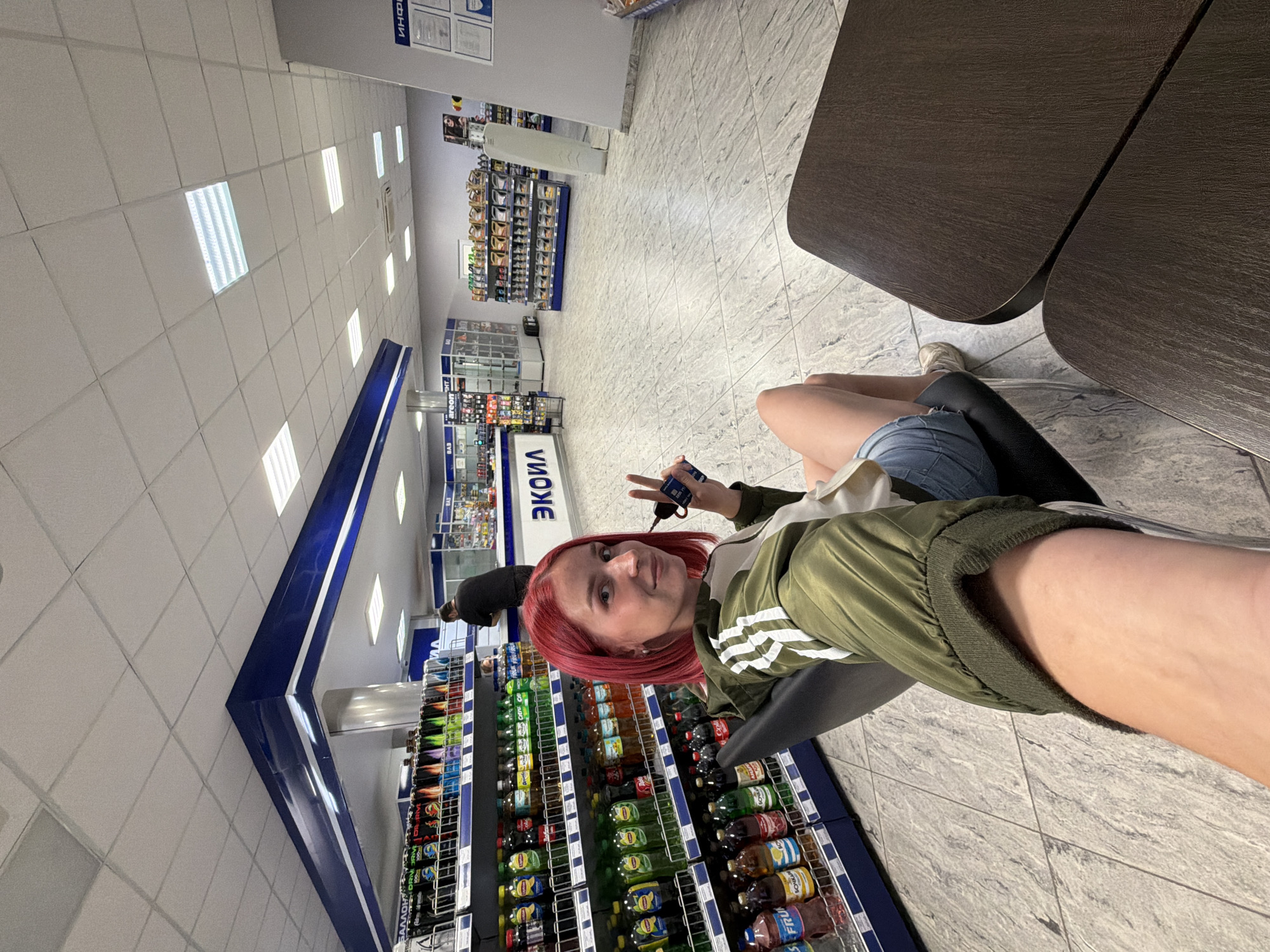 In the city of Chaikovsky, Russia, a woman with bright red hair is seated in a black chair, holding a smartphone and making a peace sign with her fingers. She is wearing a green bomber jacket with white stripes on the sleeves and denim shorts. The woman is positioned in front of shelves stocked with various bottled beverages, including sodas and juices. In the background, a man in a dark shirt is visible near a counter. The scene appears to be set in a convenience store or gas station mart, with tiled flooring and a white drop ceiling with fluorescent lighting. The store's signage displays the name "ЭКОИЛ" (EKOIL). The overall environment suggests a commercial retail space.