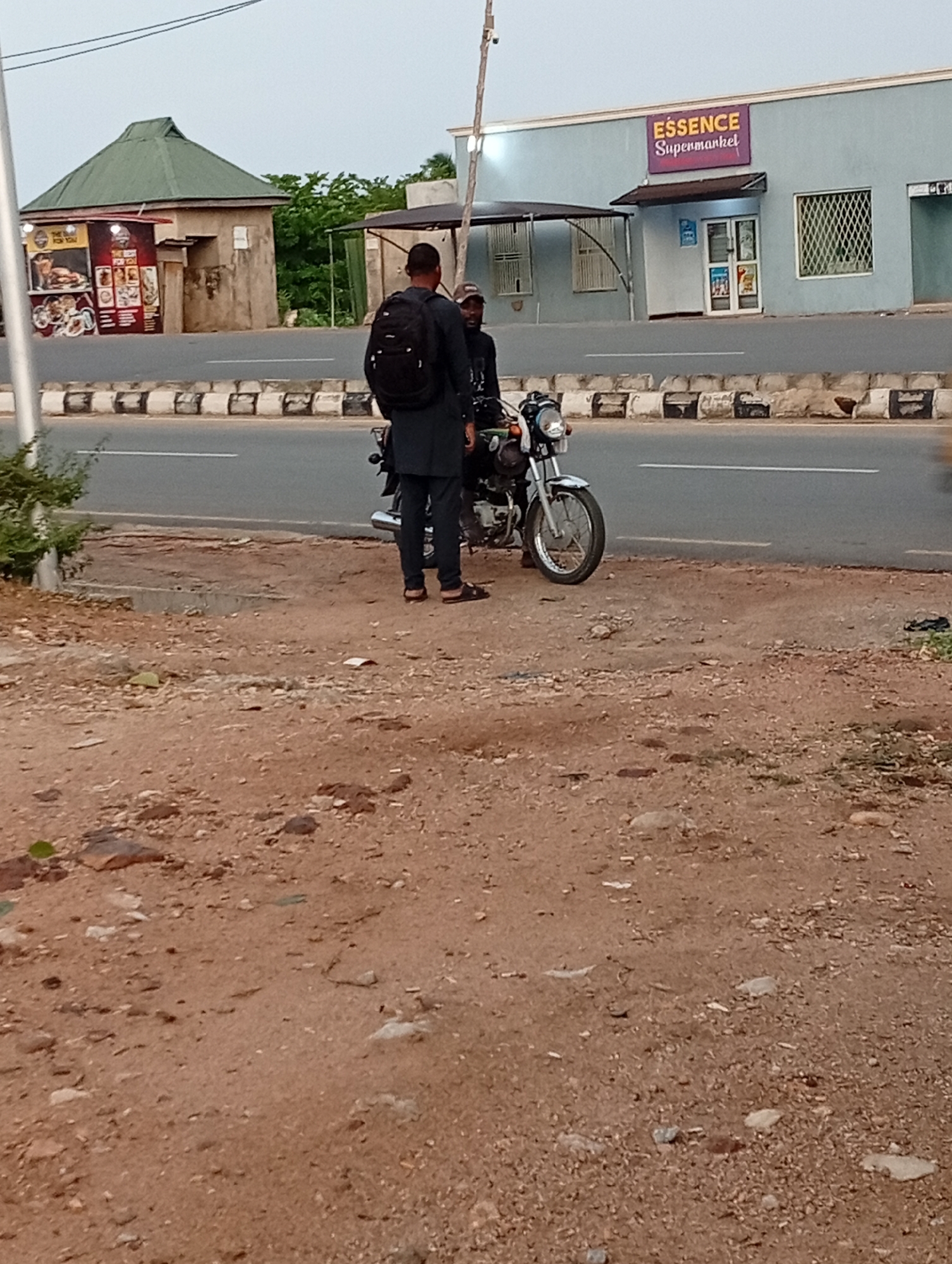 Scene: A roadside in Adankolo, Nigeria.
Subjects: Two men and a motorcycle are the primary focus. One man wears dark clothing and a backpack and stands near the motorcycle. The other man is on the motorcycle.
Objects: A motorcycle, dark backpack, and a road are prominent. In the background, there is a fast food restaurant and the "Essence Supermarket" building.
Actions: The two men appear to be interacting by the motorcycle.
Environment: A dirt and gravel shoulder is in the foreground. A road with a black-and-white curb runs in the midground. Buildings are in the background, suggesting a commercial area. The sky is overcast.
