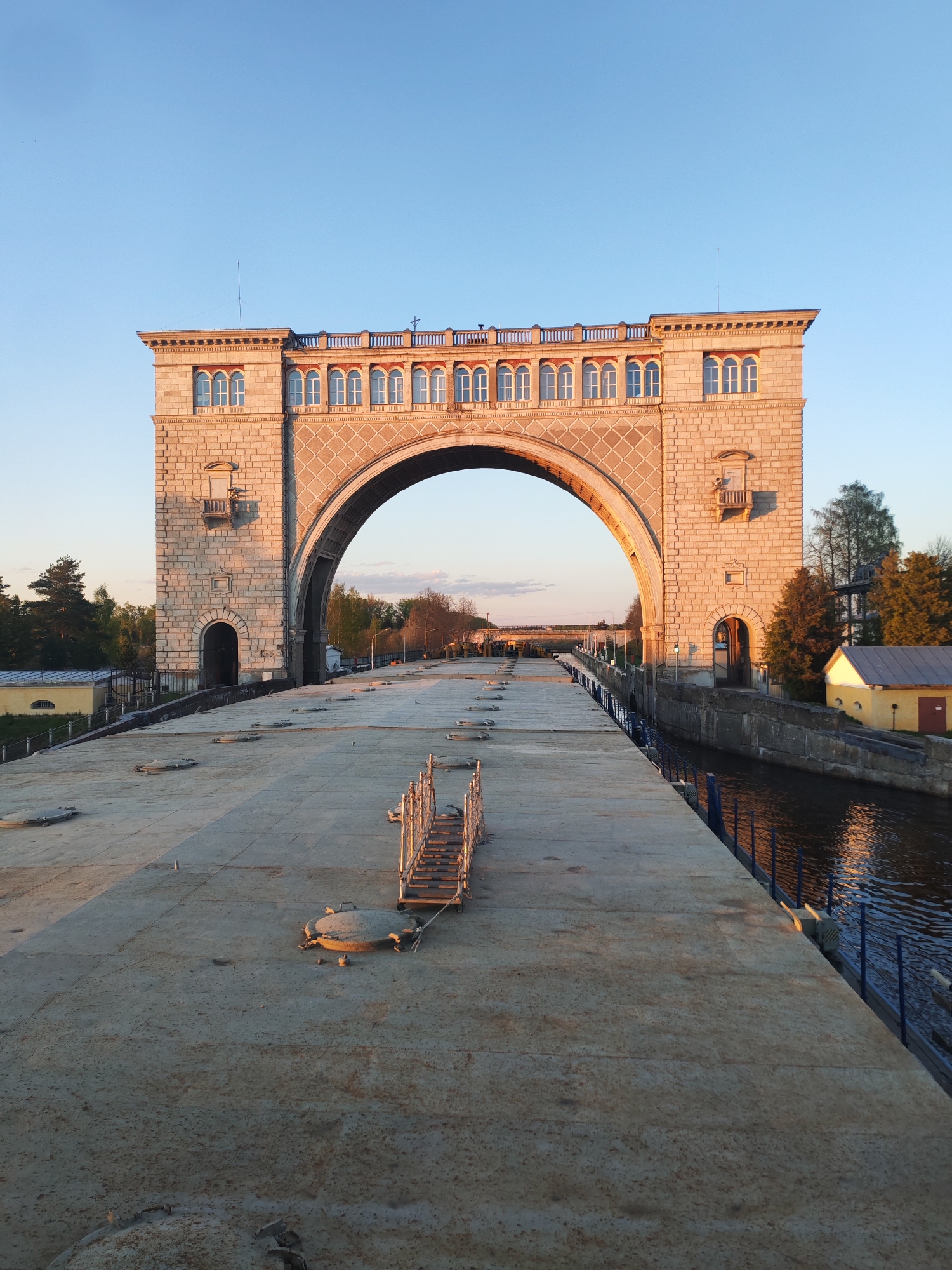 The image depicts a large, arched structure, likely a lock or gate, over a canal. The structure is built from stone and has a rectangular shape with a large arch in the center. It features windows and small balconies on both sides. The canal is flanked by concrete platforms with metal railings. In the background, there is a distant landscape with trees and a clear sky. The scene appears to be in Chelganovo, Russia. The picture was taken during sunset.