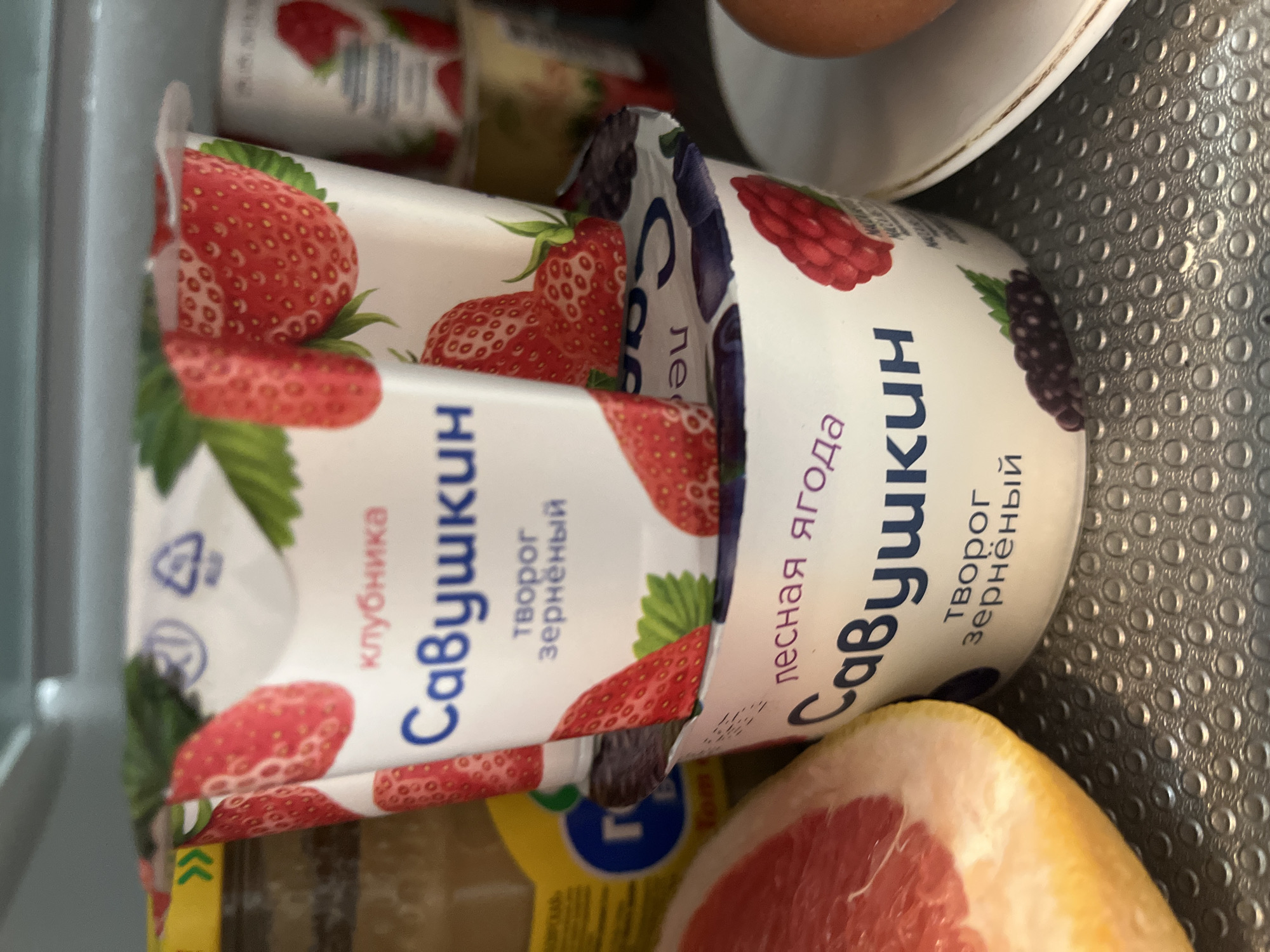 The scene captures the interior of a refrigerator, displaying various food items. In the center, two white plastic containers of "Савушкин" brand granular cottage cheese ("творог зернёный") are stacked. The upper container is strawberry-flavored ("клубника"), featuring illustrations of red strawberries and green leaves, with a partially visible lid showing recycling symbols. The lower container is forest berry-flavored ("лесная ягода"), decorated with images of red raspberries and dark berries. To the lower left, a half-cut grapefruit displays its pink flesh and yellow rind. To the lower right, a single brown egg rests on a white plate. A yellow jar with a partial blue logo is visible on the far left. The items are situated on a grey, perforated refrigerator shelf. No individuals are present, and no actions or interactions are depicted. This scene is located in Minsk, Belarus.