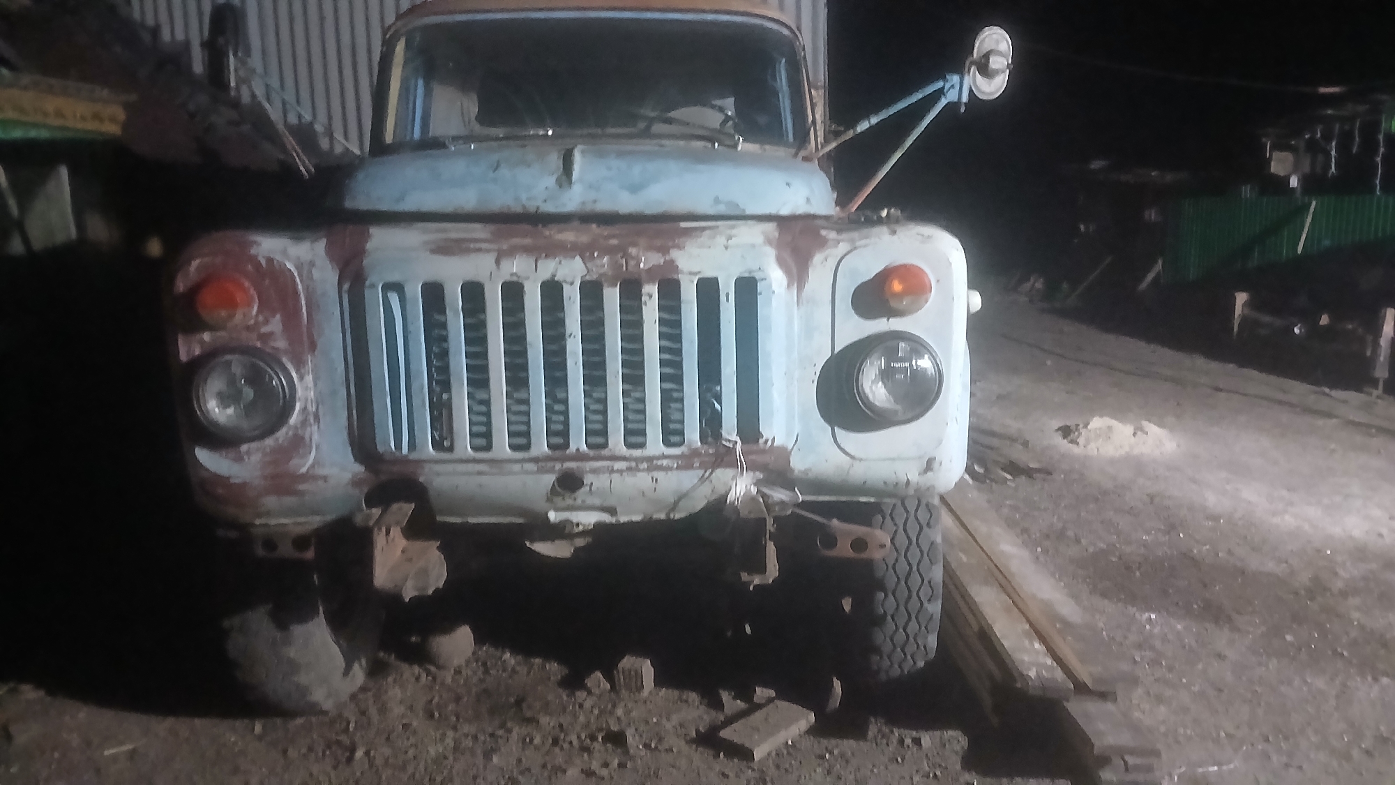 The image shows the front of a light blue, older model truck parked outdoors at night. The truck is weathered, with visible rust on the hood and fenders. It has a prominent grille, round headlights, and orange turn signals. The truck is on a dirt or gravel surface, with some scattered bricks and debris around it. In the background, there are some structures, including what appears to be a corrugated metal wall and some green fencing, suggesting an industrial or storage area. The lighting is dim, typical of a nighttime scene, with a light source illuminating the front of the truck. There are no people visible in the image. The location is identified as Slak, Russia.