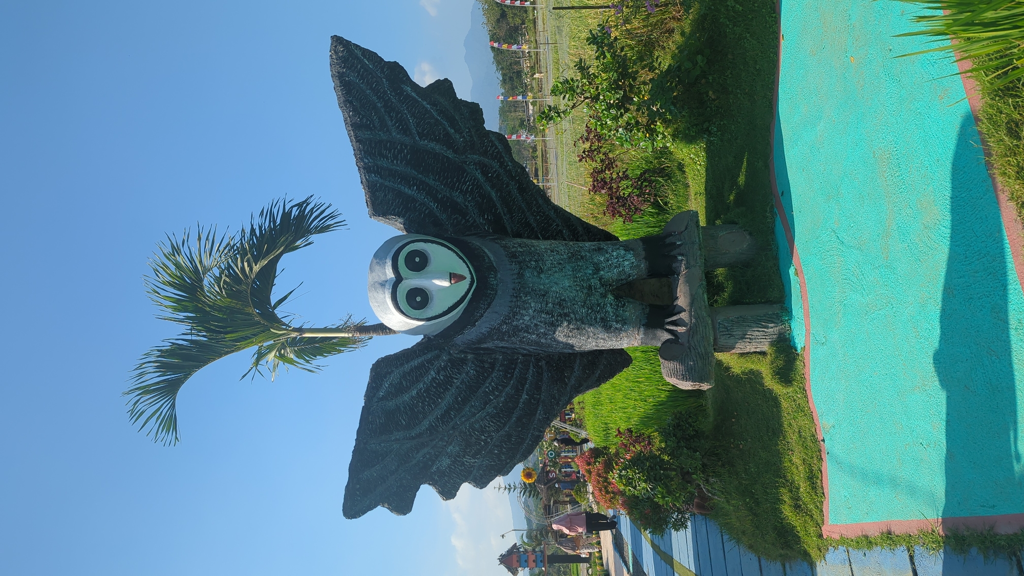 A full-frame, vertical, eye-level outdoor shot features a large owl statue with outstretched wings at the center of the frame. The statue is dark gray with a white face featuring large black eyes and a small red beak. It stands on a wooden log base, which is supported by two smaller logs. Behind the owl, a bright green palm tree with a curved trunk and fronds reaches towards a clear, bright blue sky.
To the left of the owl, a blue and yellow pathway winds through a grassy area with red-leafed bushes. In the background, several people, one wearing a pink headscarf, walk along the path. Further behind them are small structures, including a birdhouse-like building and a tall yellow sunflower. The ground beyond the immediate grassy area is a vibrant green, appearing to be a rice paddy.
The lower portion of the image shows a bright turquoise-colored surface with a reddish-pink border, possibly a decorative ground covering or a shallow water feature. Tall blades of green grass are visible in the bottom right corner. In the far distance, a mountain range is visible, and several colorful flags are planted in a field. The overall scene suggests an outdoor recreational area or park. The location is identified as a tourist attraction in Semarang, Indonesia, specifically the "Wisata Agro Kampoeng Djowo Sekar" or "Kampoeng Djowo Sekar Agro Tourism," known for its scenic landscapes and photo spots.